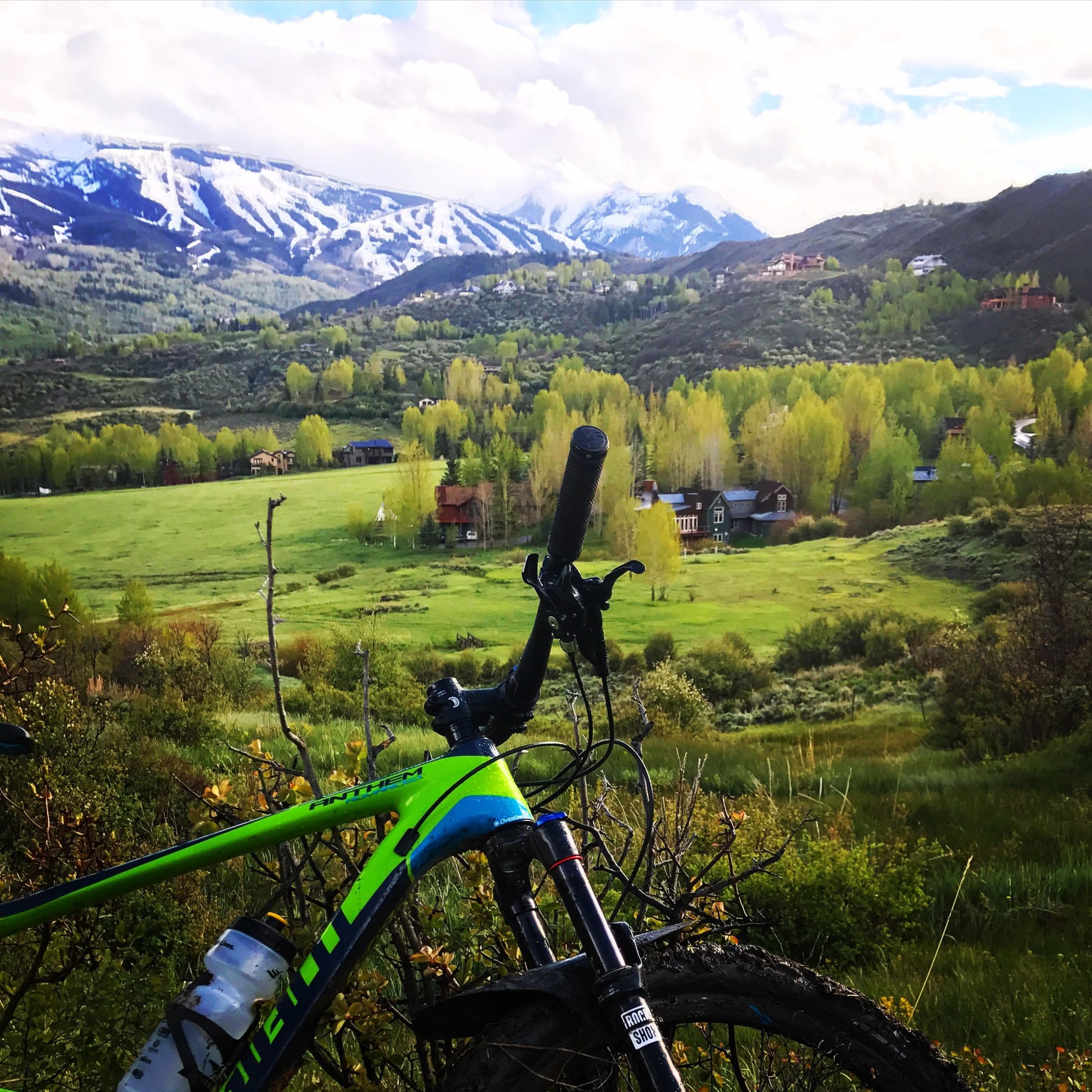 A vibrant mountain landscape in spring featuring a green and black mountain bike in the foreground, with a backdrop of rolling hills, lush green fields, and scattered houses. Snow-capped mountains rise majestically in the distance under a partly cloudy sky. Rim Trail mountain bike trail.