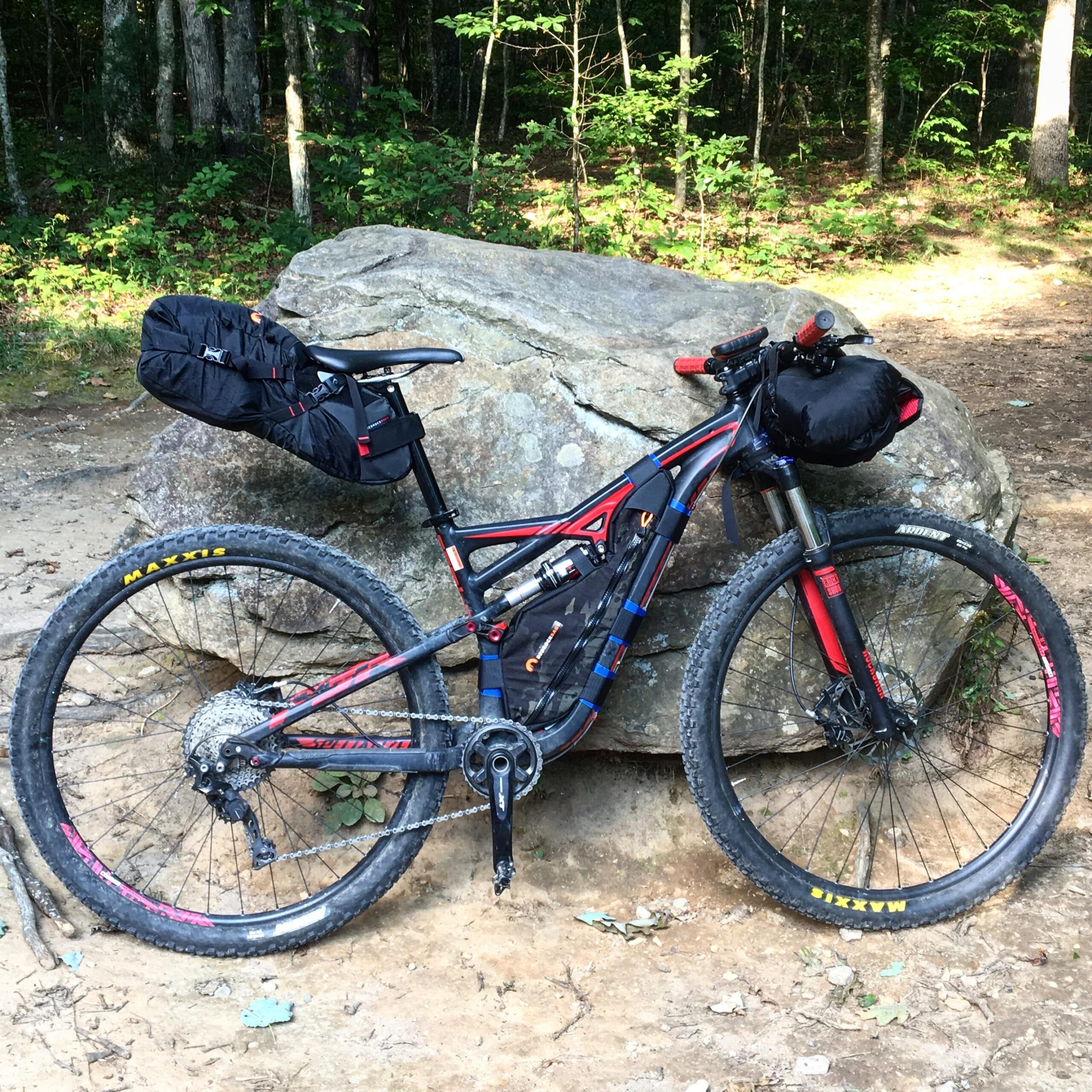 Specialized Camber 29: A mountain bike with black and red accents is leaning against a large rock in a wooded area. The bike features a frame bag and additional gear attached to the seat and handlebars. The scene includes lush green trees in the background and a dirt trail, suggesting an outdoor adventure setting.