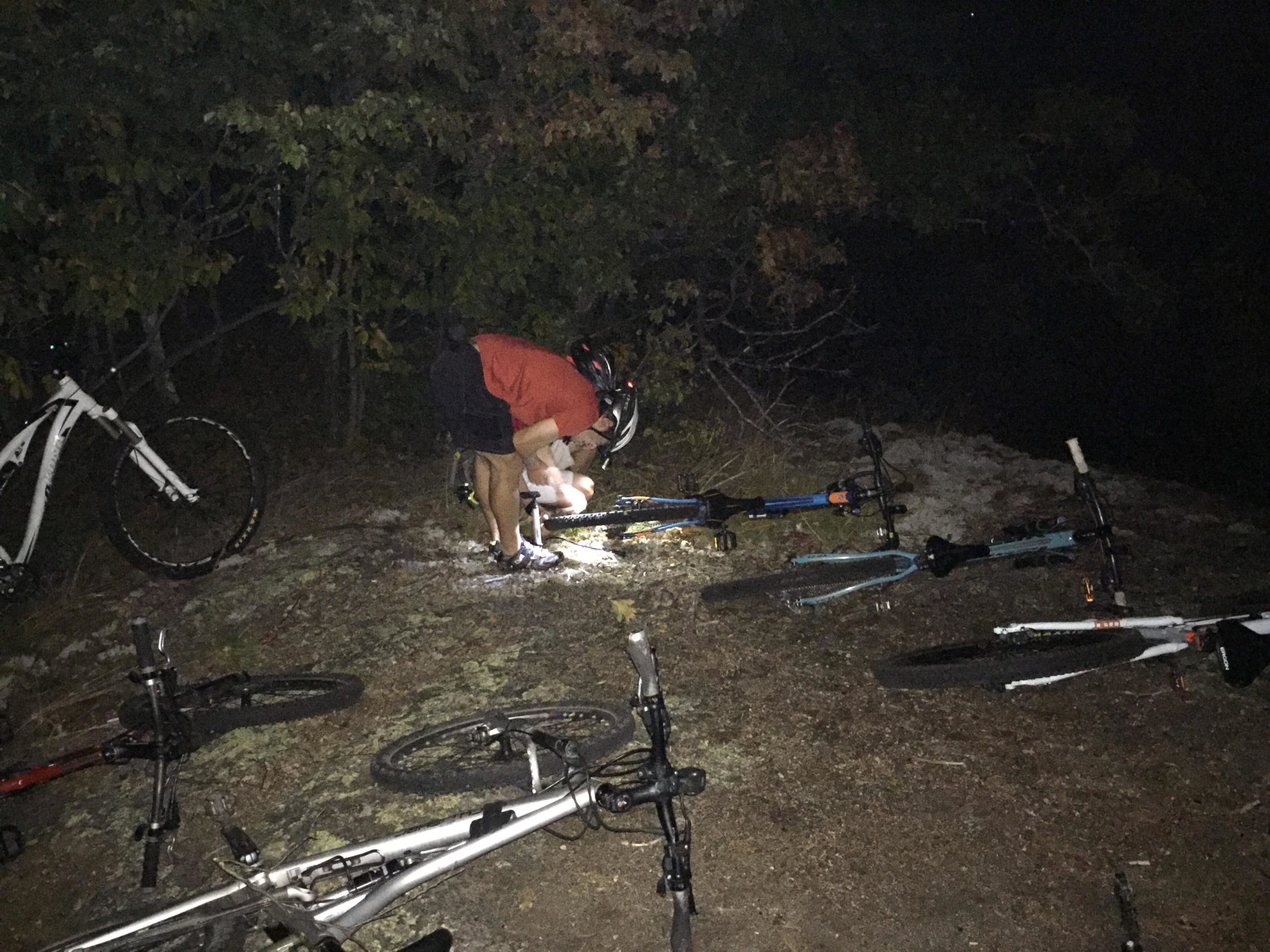 A person in a red shirt bends down to work on a mountain bike, using a flashlight to illuminate the area at night. Several other bikes lay on the ground nearby, surrounded by trees and foliage. Blueberry Hill mountain bike trail.