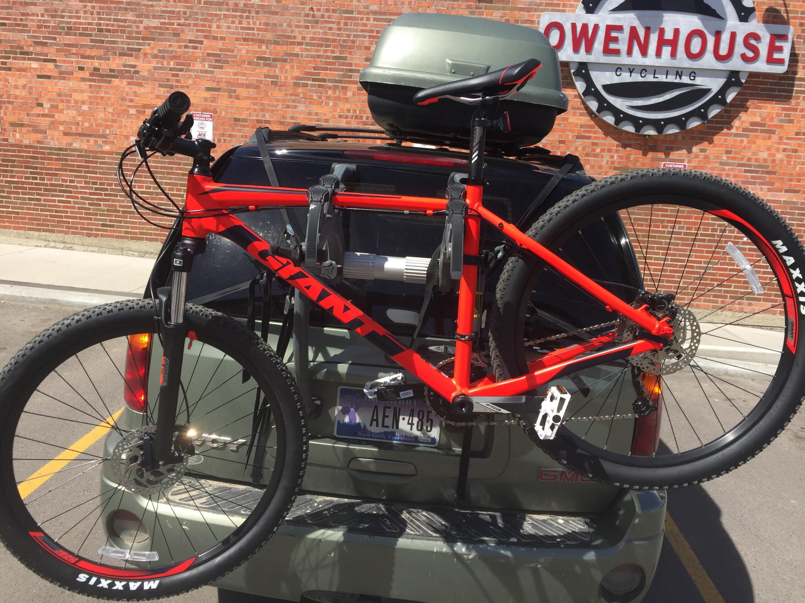 Giant Talon 27.5 2: A red Giant mountain bike securely mounted on a bike rack attached to the back of an SUV. The vehicle is parked in front of a brick wall with a sign for Owenhouse Cycling in the background. The bike features thick tires designed for off-road riding.
