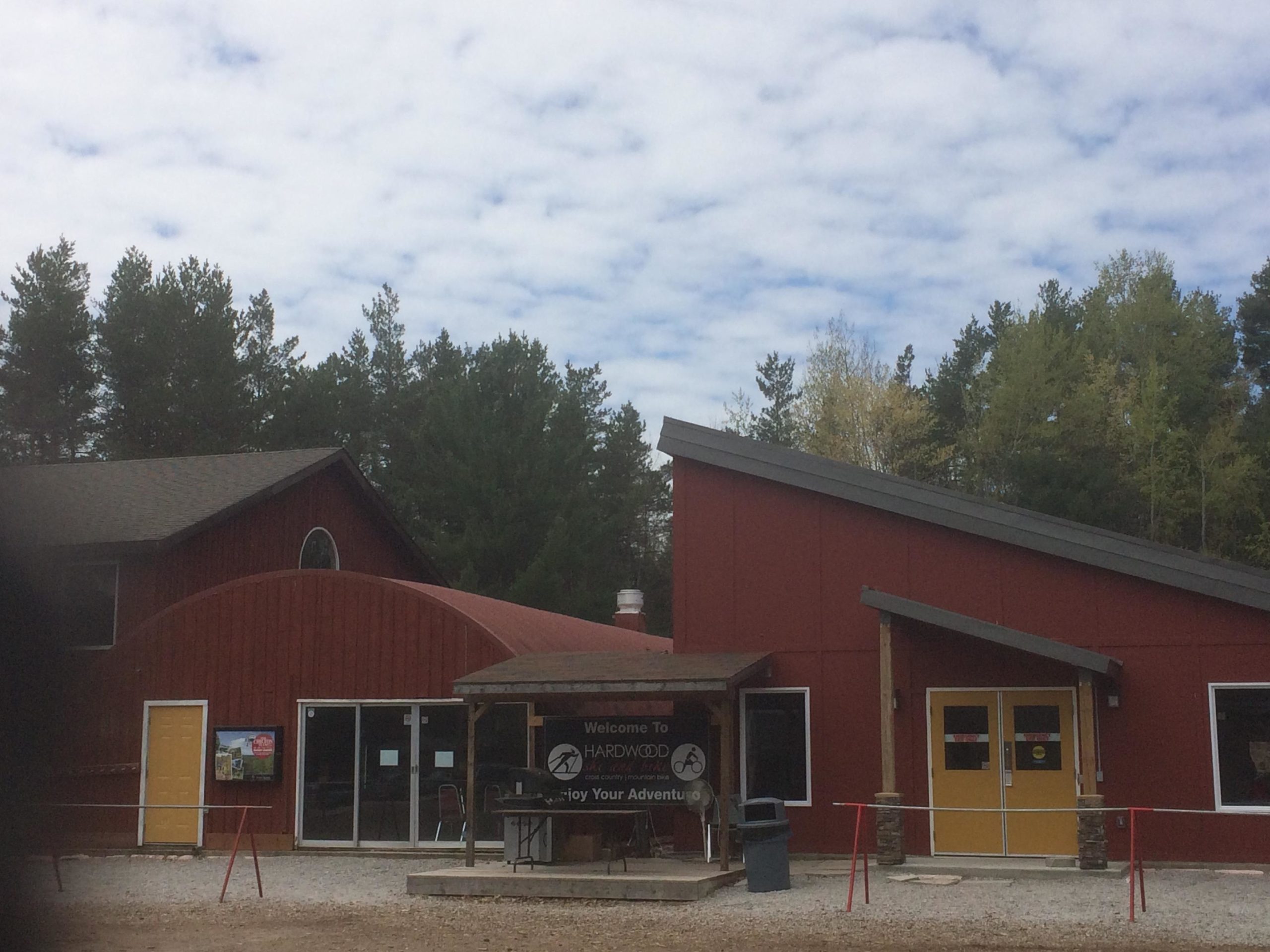 A rustic, red building with a unique architecture featuring an arched roof and sharp angles, set against a backdrop of green pine trees under a partly cloudy sky. The entrance includes a welcoming sign and yellow doors, with a gathering area visible in front. Hardwood Ski and Bike mountain bike trail.