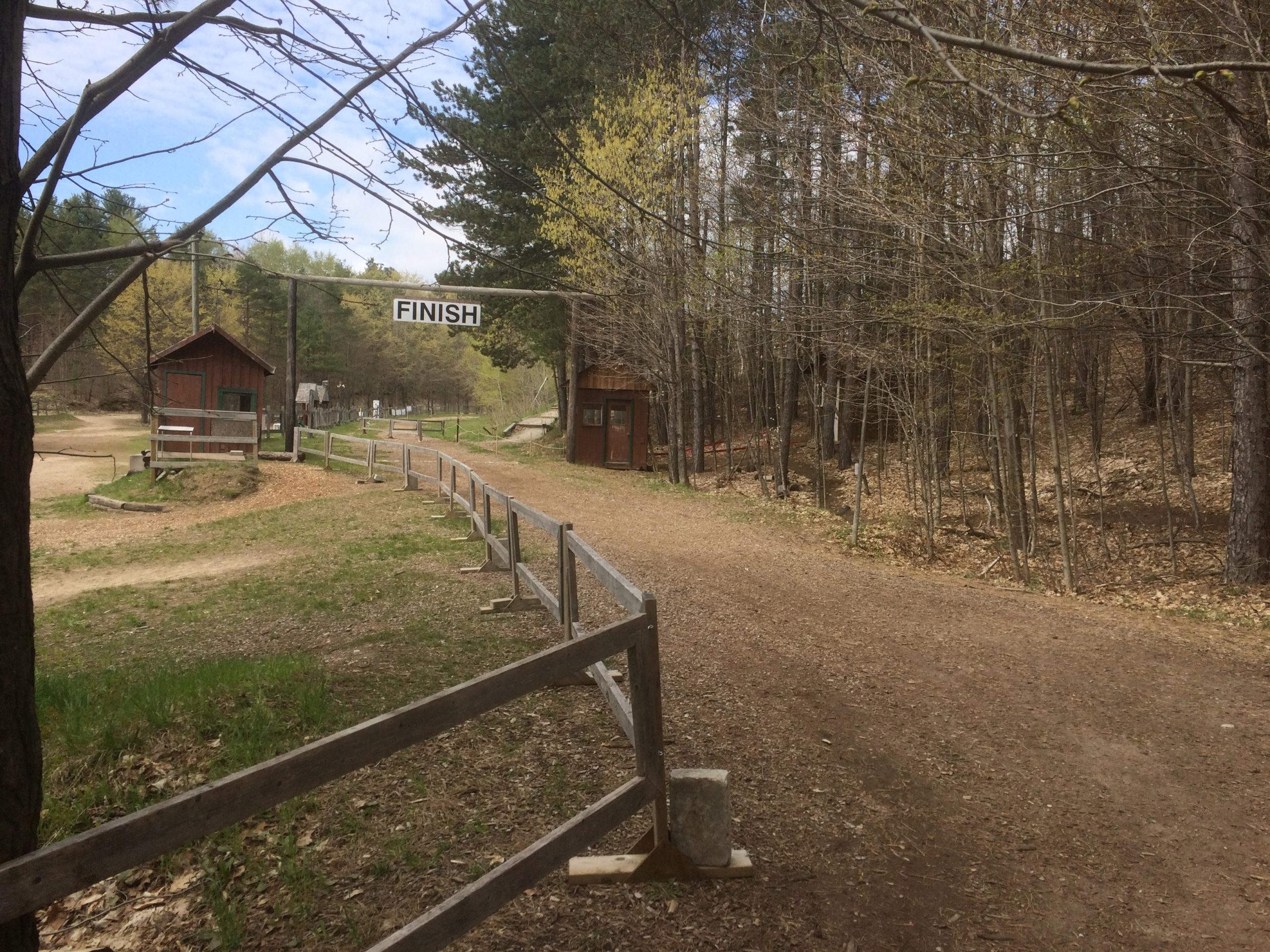 A dirt path leading to a "FINISH" sign, surrounded by trees and wooden structures. The scene showcases a wooded area with a clear sky and signs of early spring. Hardwood Ski and Bike mountain bike trail.