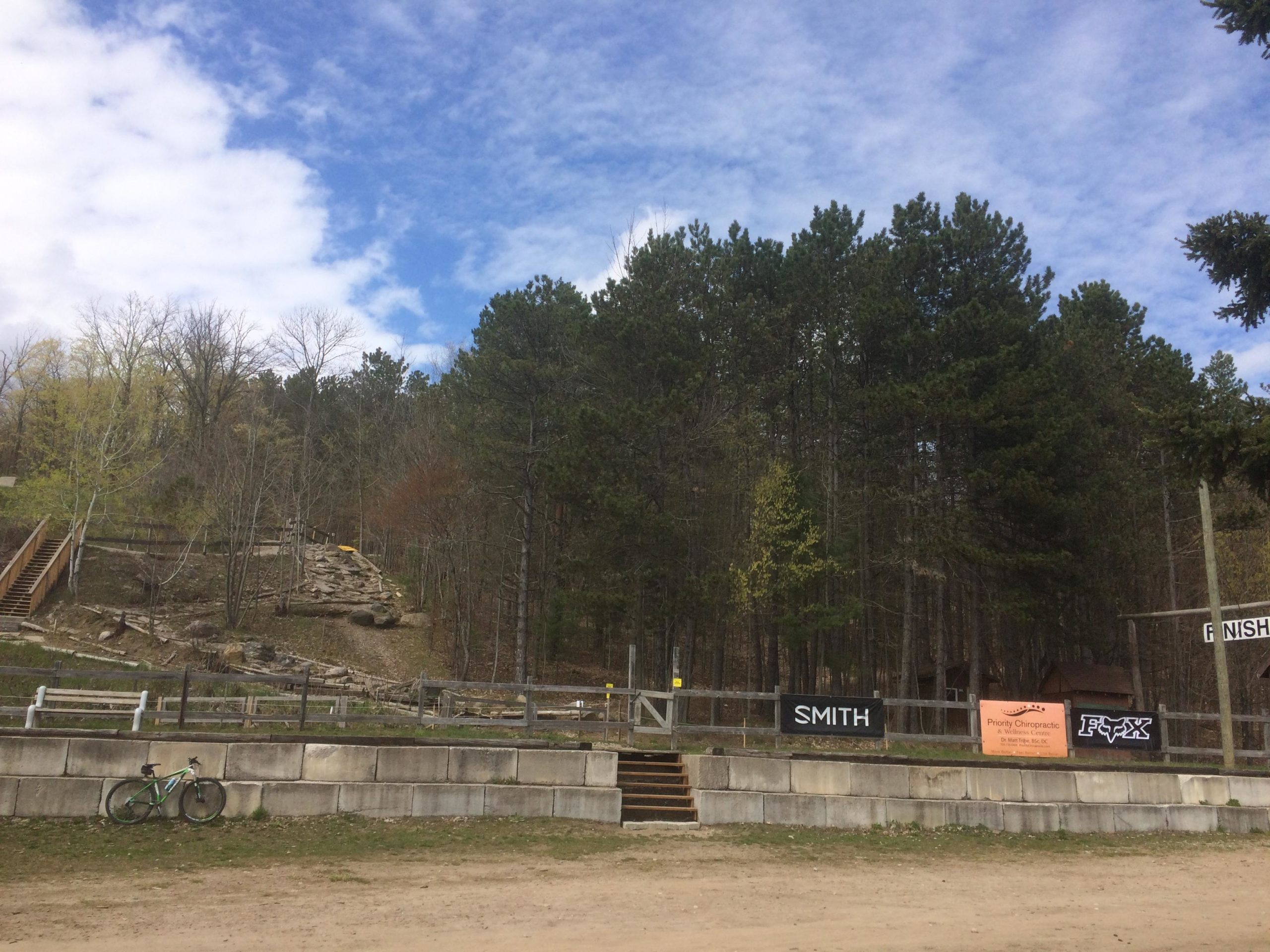 A dirt trail leading to a forested area with tall pine trees under a partly cloudy sky. In the foreground, a green bicycle leans against a stone wall, and there are stairs ascending the hill. Several banners for different sponsors are displayed along a wooden fence. Hardwood Ski and Bike mountain bike trail.
