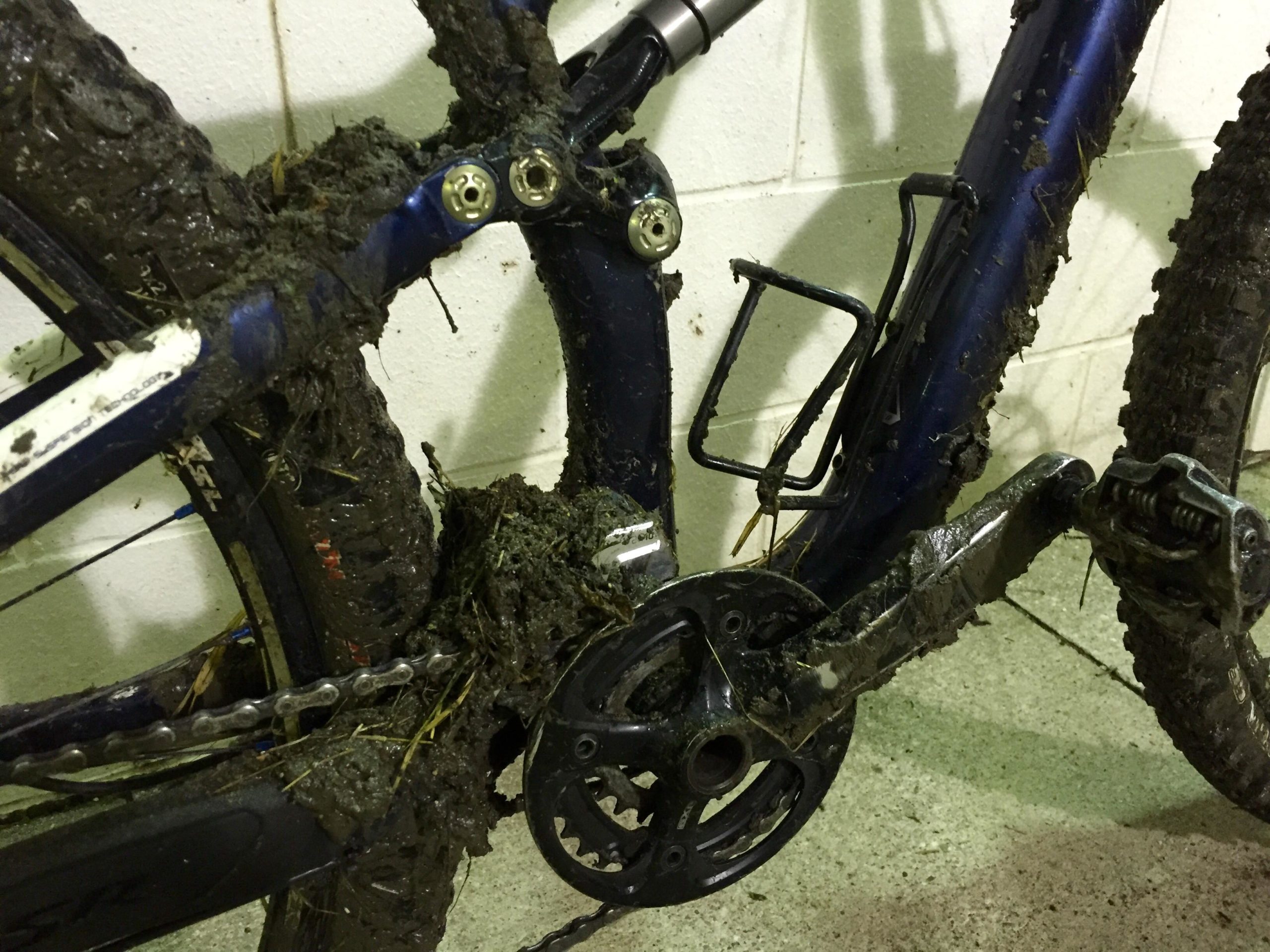 A close-up image of a muddy mountain bike, highlighting the heavily soiled frame, chain, and drivetrain components. The bike is leaning against a wall with a plain white background, showcasing the accumulation of mud and grass from off-road riding. Angel Fire Bike Park mountain bike trail.