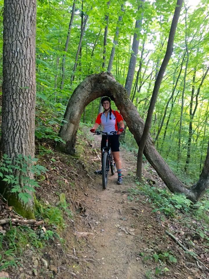 A person on a mountain bike poses next to a uniquely shaped tree arch on a forest trail, surrounded by lush green foliage. The individual is wearing a helmet and a bright shirt, smiling while the bike is parked beside them. The Bluff / Missing Link mountain bike trail.