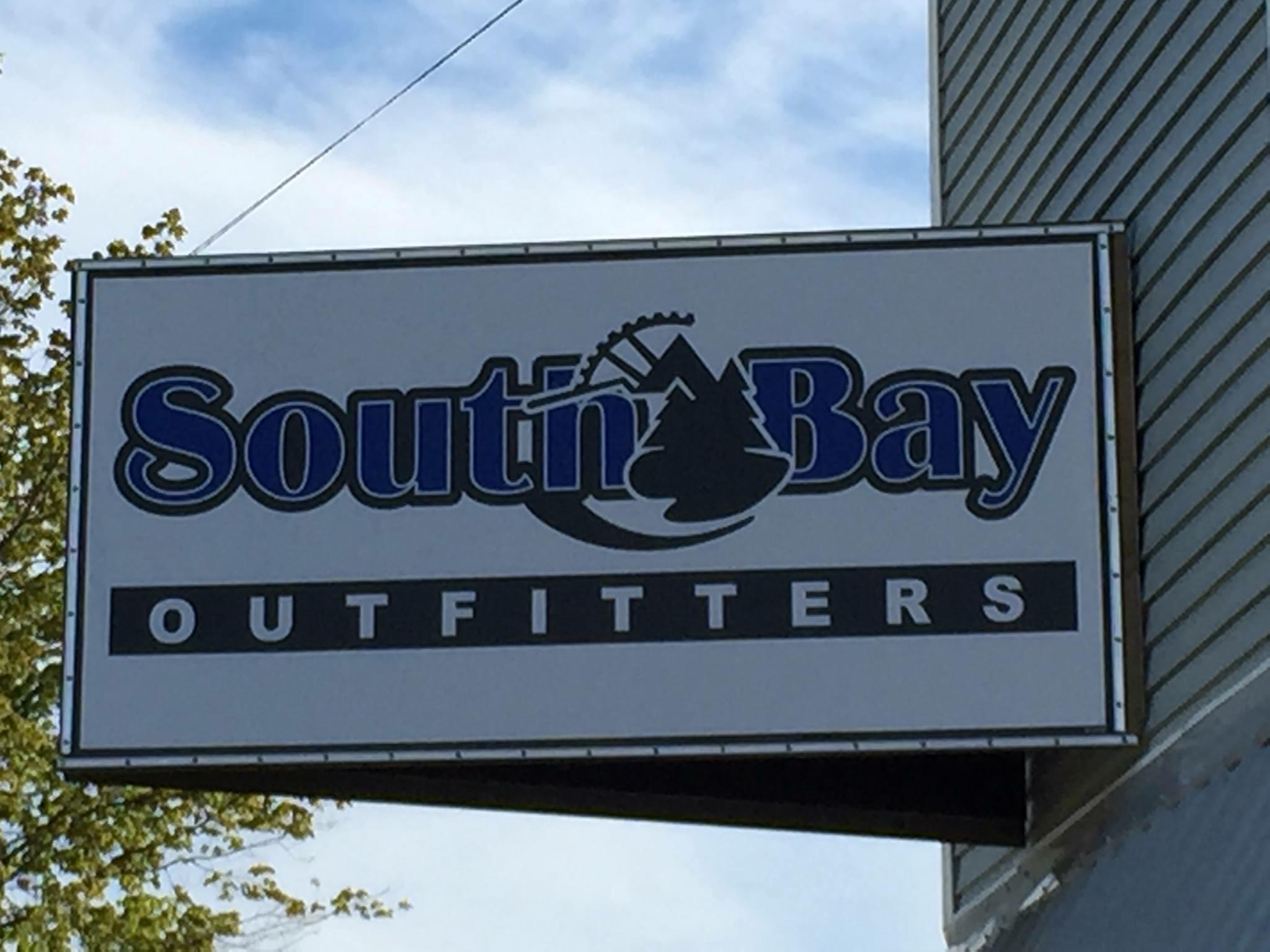 Sign for South Bay Outfitters, featuring the name prominently in blue and black lettering, accompanied by imagery of trees and mountains. The background shows a partly cloudy sky.