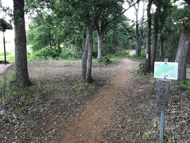 Pathway through a wooded area with trees on either side, leading to a sign indicating a golf course nearby. The ground is covered with dirt and scattered leaves, and a small map of the course is visible on the signpost. Horseshoe mountain bike trail.