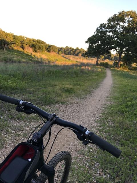 A mountain bike resting on gravel trail, surrounded by lush green fields and trees, with a scenic path winding through the landscape under a clear sky. Horseshoe mountain bike trail.