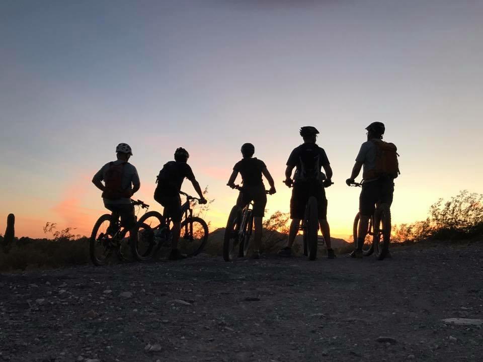Silhouette of five mountain bikers standing on a dirt path, facing a sunset with colorful skies. The riders are equipped with helmets and bikes, surrounded by desert vegetation. 40th St. Trailhead mountain bike trail.