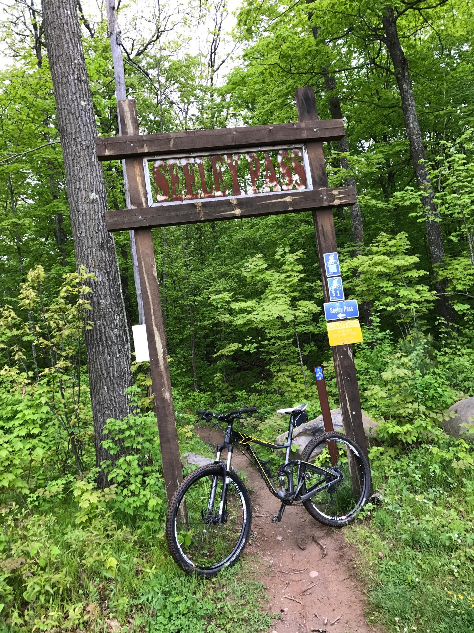 A wooden sign displaying "Seeley Pass" marks the entrance to a mountain biking trail surrounded by lush green foliage. A black mountain bike rests against the sign, with a pathway leading into the forest. Additional signage indicates trail information and a caution warning. Seeley Pass Trail mountain bike trail.