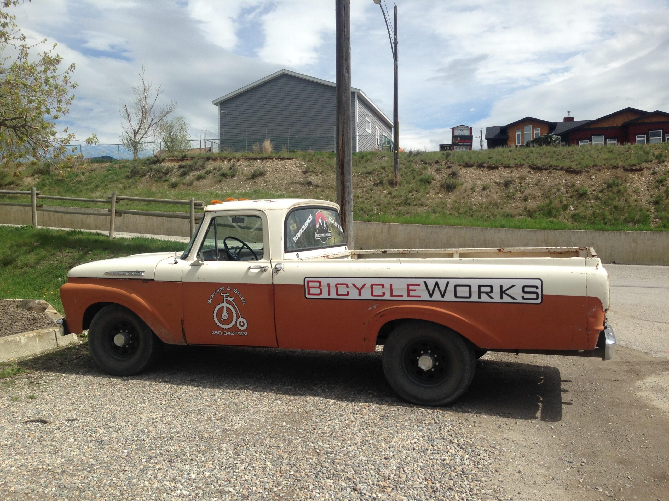 A vintage pickup truck parked on a gravel surface, featuring a two-tone design with a white cab and orange body. The side of the truck displays the logo and text for "Bicycle Works," indicating it is associated with bicycle service and sales. In the background, there are grassy hills and a wooden fence, along with modern buildings. The sky is partly cloudy.