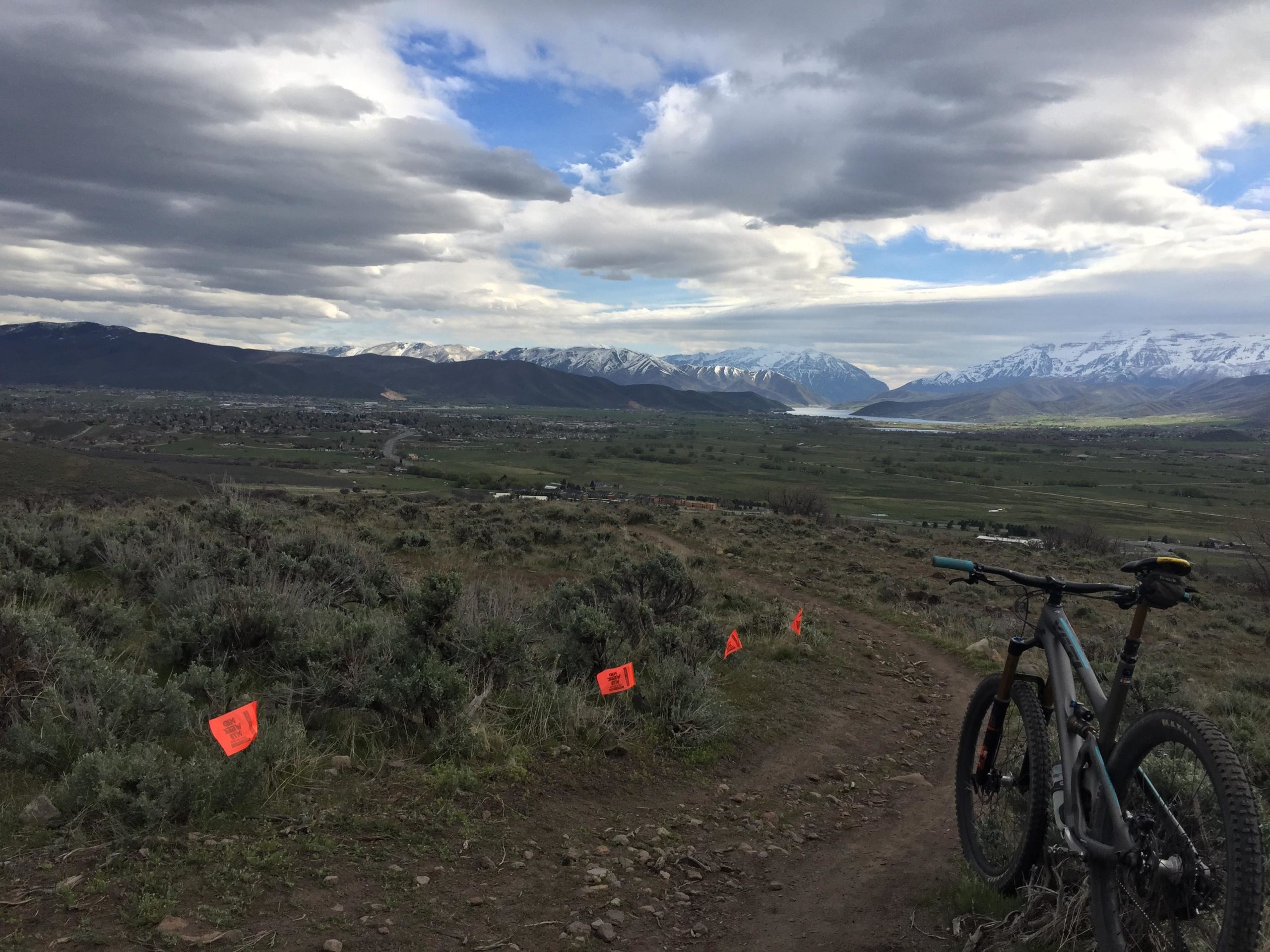A mountain bike resting on a dirt trail overlooking a scenic valley. In the background, snow-capped mountains rise under a cloudy sky, with patches of greenery and orange flags marking the terrain below. Chopped Coyote Loop mountain bike trail.