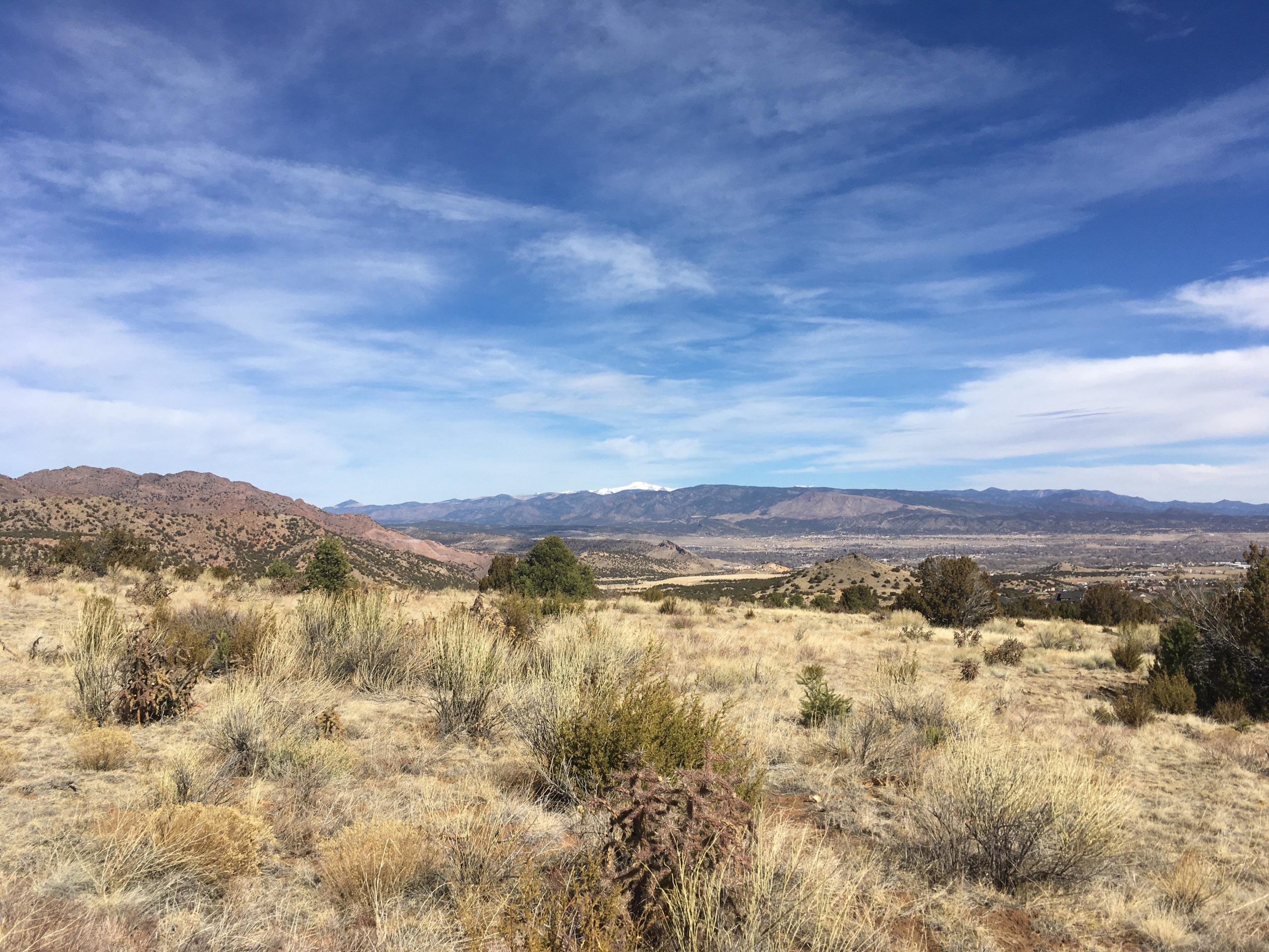 A panoramic view of a dry, mountainous landscape with rolling hills and sparse vegetation. The foreground features grasses and small shrubs, while distant mountains are seen under a bright blue sky with wispy clouds. Snow-capped peaks are visible in the far distance, suggesting a varied terrain. South Canon Trails mountain bike trail.