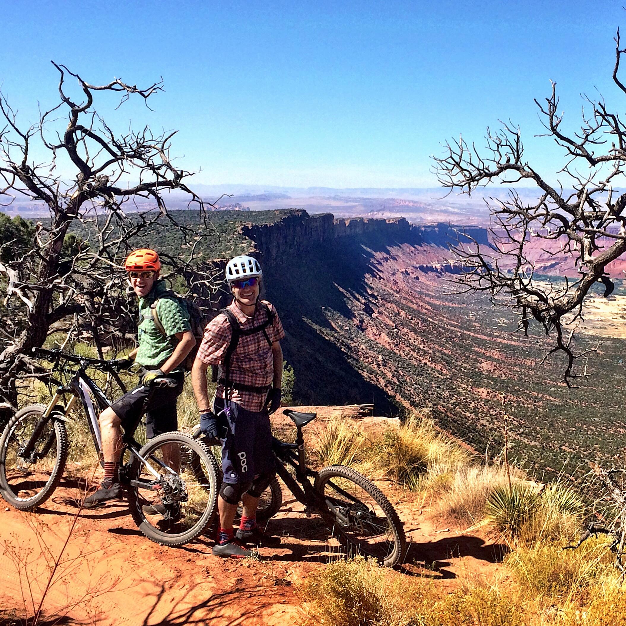 Specialized Epic Expert Carbon 29er: Two mountain bikers standing on a rocky overlook with scenic views of a canyon and rugged terrain in the background. One biker is wearing an orange helmet and green shirt, while the other is in a white helmet and plaid shirt. The landscape features dry vegetation and distant cliffs under a blue sky.