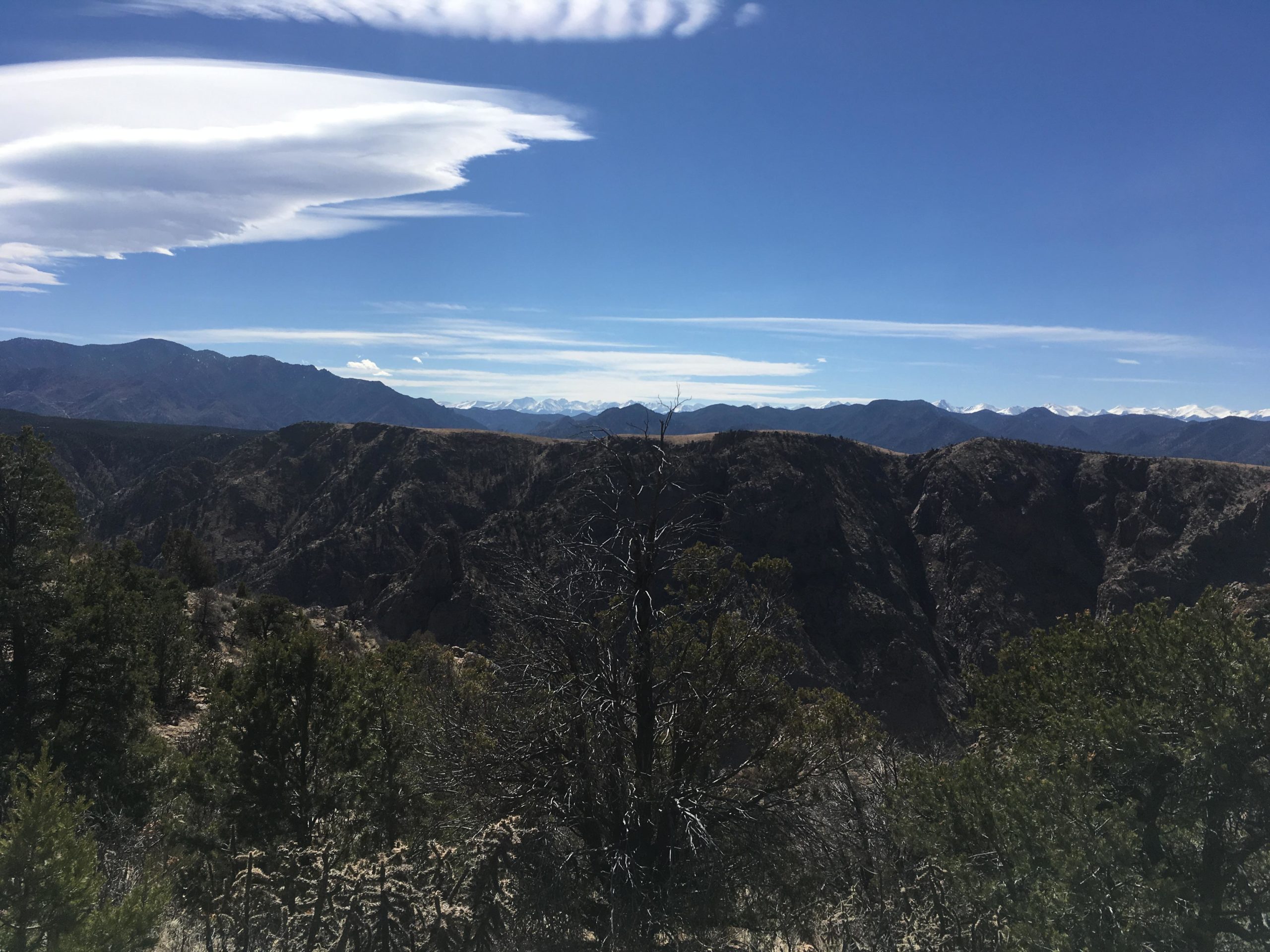 A panoramic view of rugged mountain terrain under a clear blue sky, featuring elevated peaks in the distance and unique cloud formations. The landscape includes rocky cliffs and patches of greenery, with a contrasting backdrop of snow-capped mountains on the horizon. Royal Gorge Park Trail System mountain bike trail.