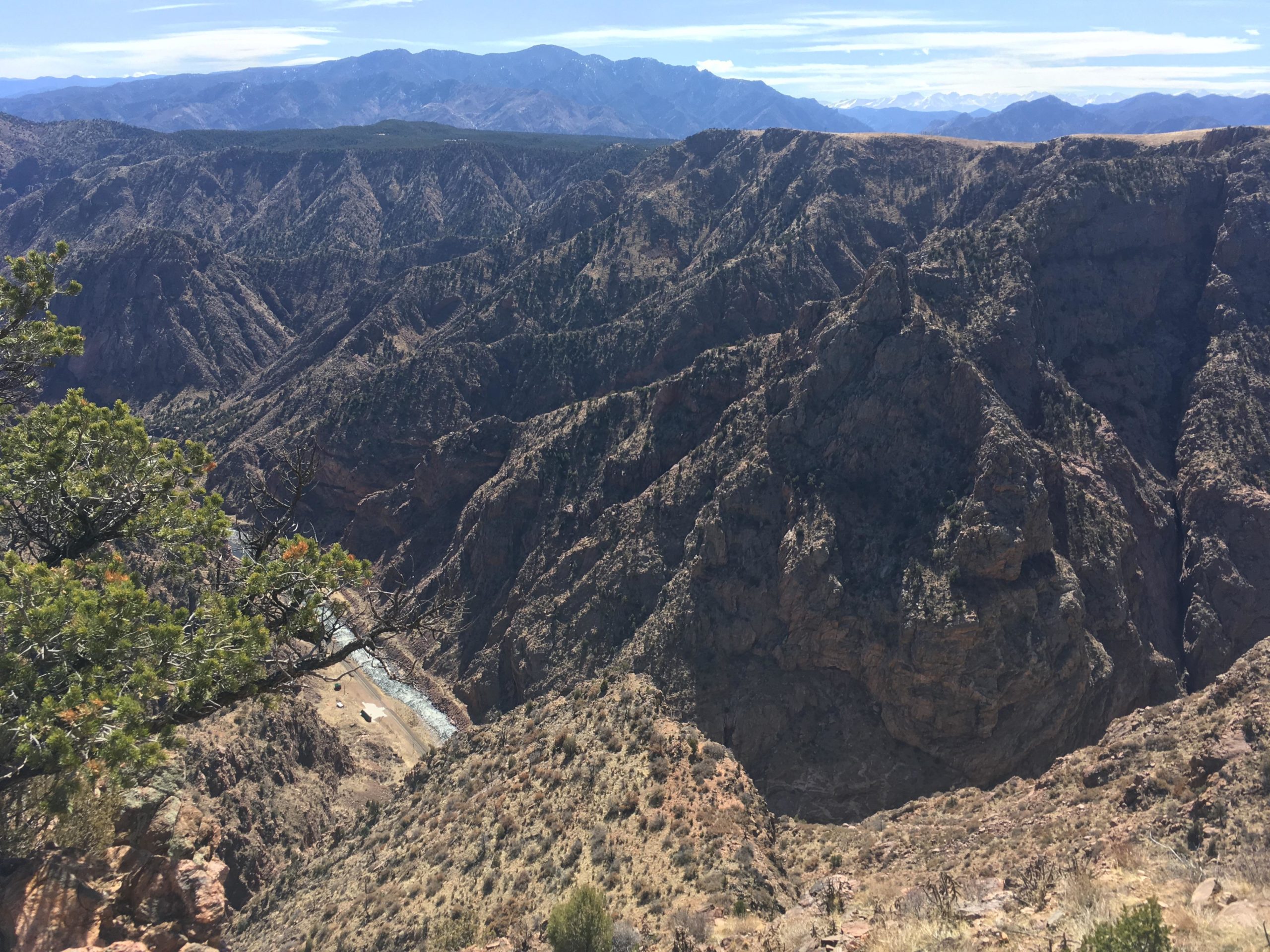 A breathtaking landscape featuring rugged mountain ridges and deep valleys, with a river winding through the terrain. The scene is bathed in bright sunlight under a clear blue sky, showcasing the natural beauty of the rocky landscape and sparse vegetation. Royal Gorge Park Trail System mountain bike trail.