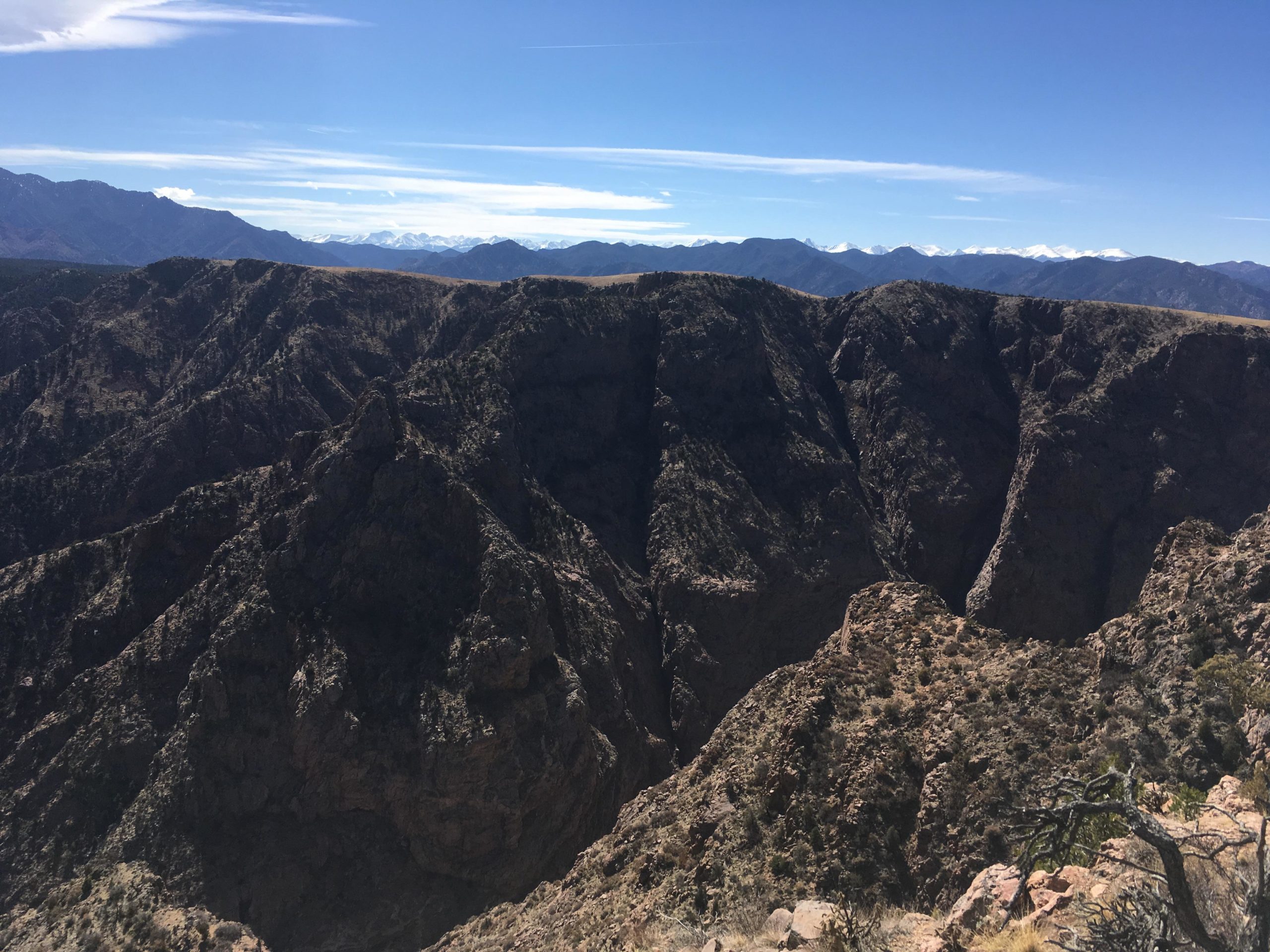 A panoramic view of rugged mountain terrain with steep cliffs and rocky formations. The landscape features deep canyons and distant snow-capped peaks under a clear blue sky with wispy clouds. The foreground includes dry vegetation and sparse trees, highlighting the natural beauty of the mountainous region. Royal Gorge Park Trail System mountain bike trail.