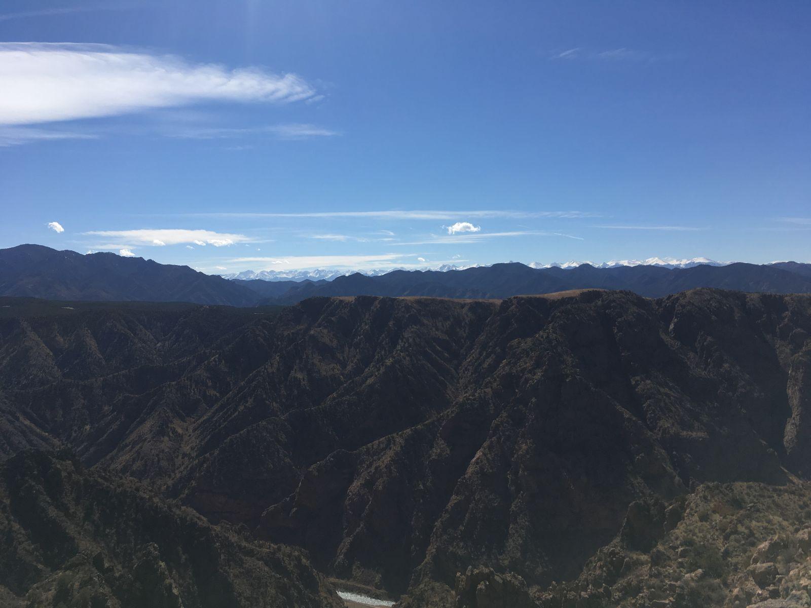 A panoramic view of rugged mountain ranges under a clear blue sky, with distant snow-capped peaks visible on the horizon. The foreground features steep, rocky slopes and dense vegetation, illustrating the natural beauty of the landscape. Royal Gorge Park Trail System mountain bike trail.