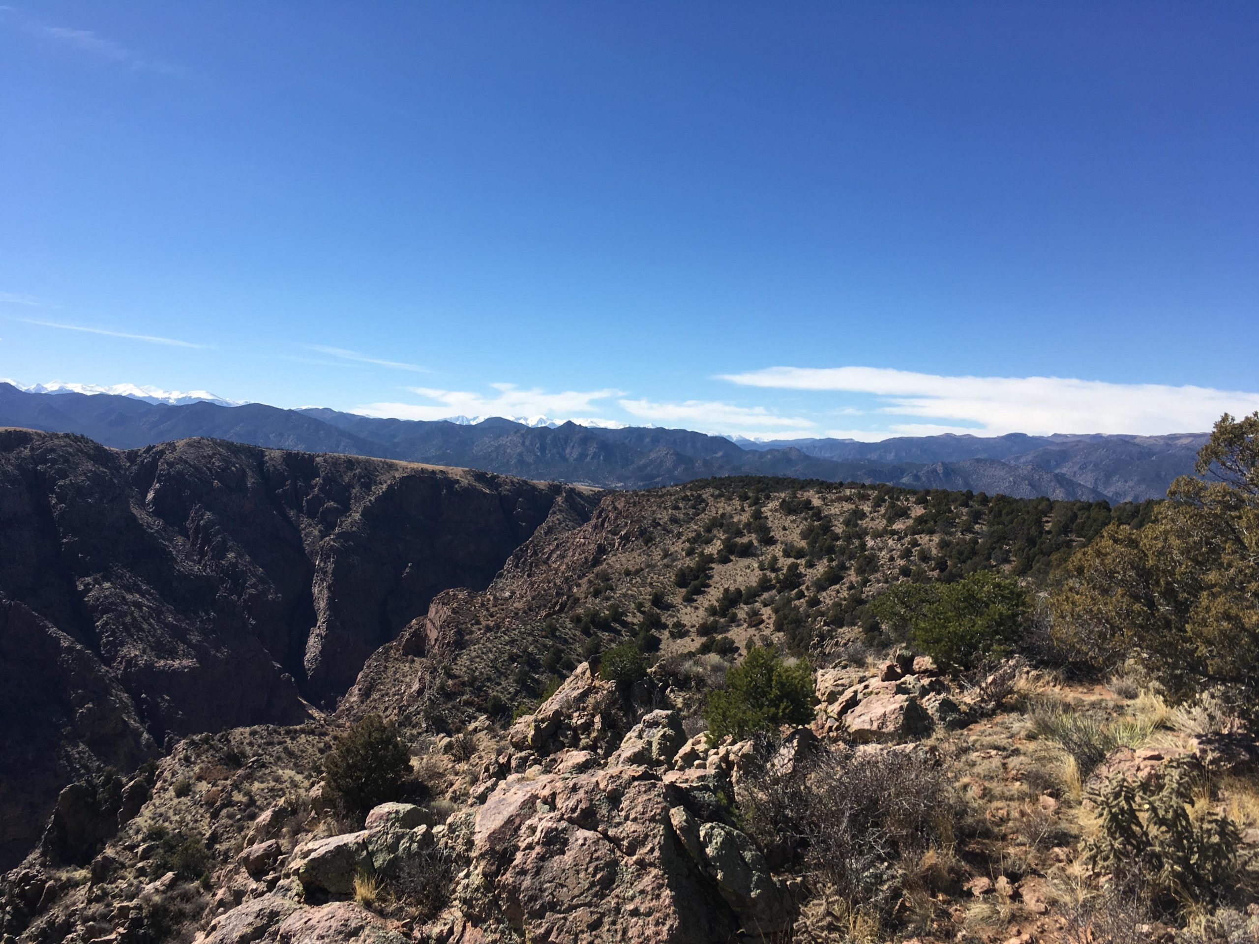 Scenic view of a mountainous landscape under a bright blue sky, featuring rocky outcrops, rolling hills, and distant snow-capped peaks in the background. Sparse vegetation and shrubs are visible in the foreground, showcasing the rugged terrain. Royal Gorge Park Trail System mountain bike trail.