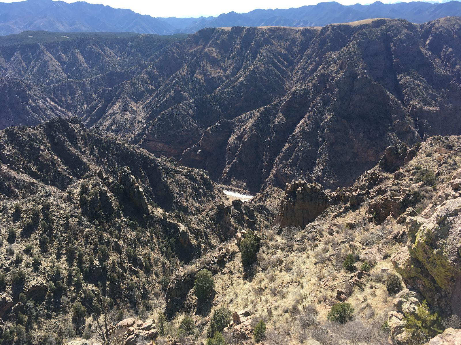 A panoramic view of rugged mountains and deep canyons under a clear blue sky, showcasing a natural landscape with rocky formations, sparse vegetation, and distant peaks in the background. Royal Gorge Park Trail System mountain bike trail.