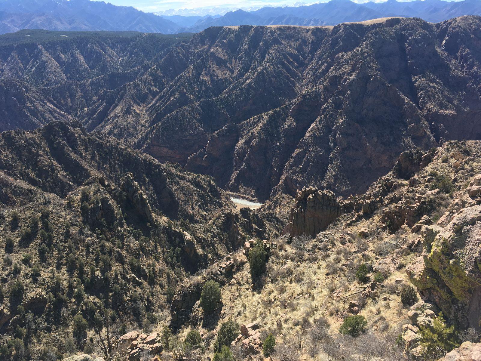 A panoramic view of rugged mountains and valleys, showcasing steep, rocky terrain covered with sparse vegetation under a clear blue sky. The scene captures the natural beauty of a remote wilderness area, with layers of mountain ridges in the background fading into the distance. Royal Gorge Park Trail System mountain bike trail.