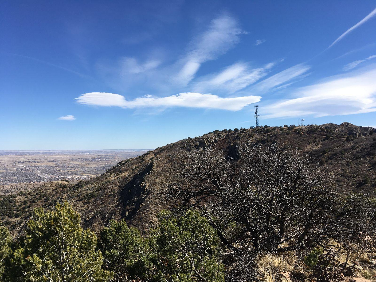 A panoramic view of a mountainous landscape featuring rugged terrain, sparse vegetation, and a clear blue sky with wispy clouds. The foreground includes some shrubbery and dry trees, while a communications tower can be seen at the summit of the hill in the background. Royal Gorge Park Trail System mountain bike trail.