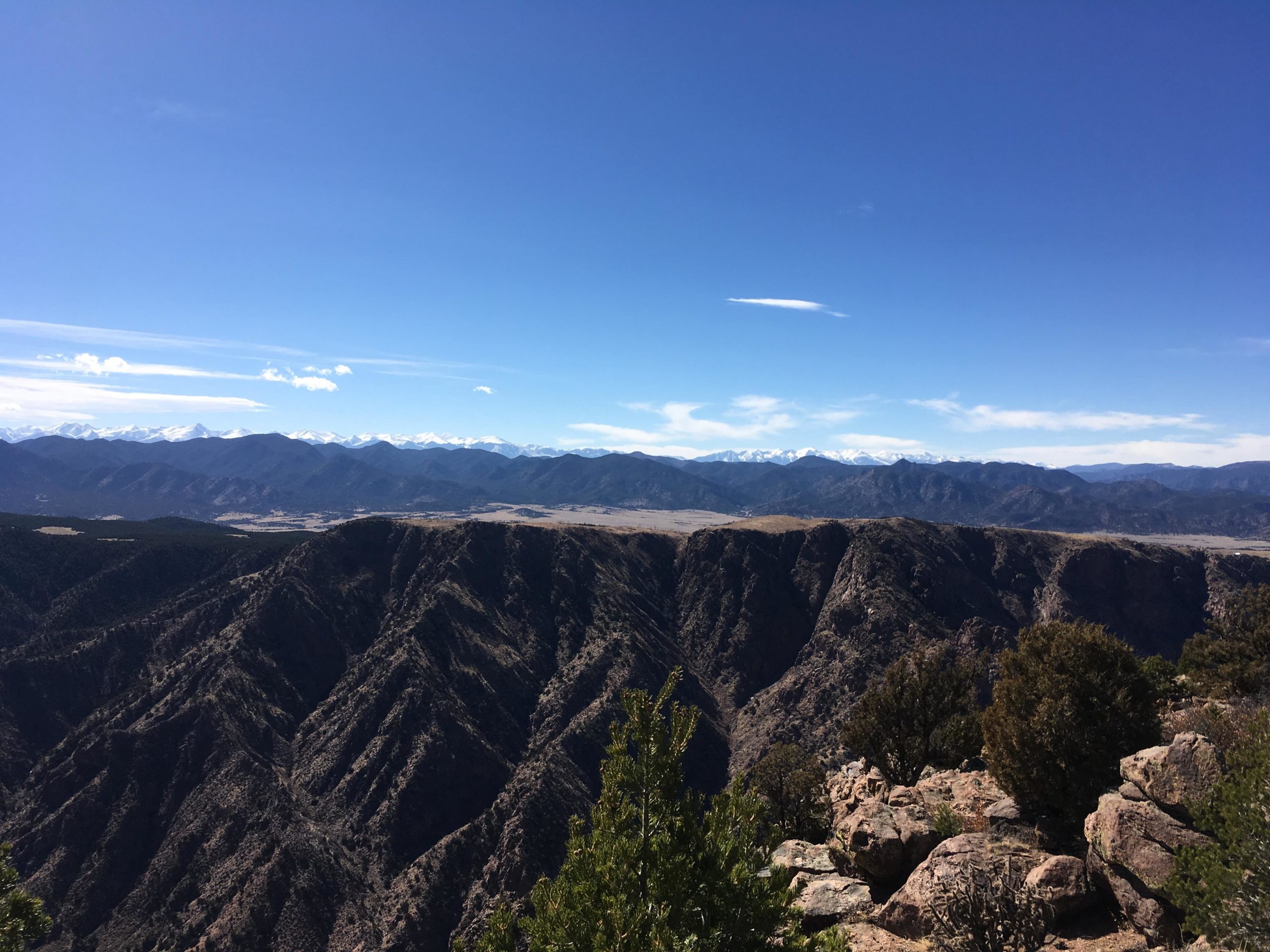 A panoramic view of rugged mountain ranges under a clear blue sky, featuring snow-capped peaks in the distance and deep valleys with rocky terrain in the foreground. Lush greenery is scattered among the rocky landscape, creating a stark contrast against the mountains. Royal Gorge Park Trail System mountain bike trail.
