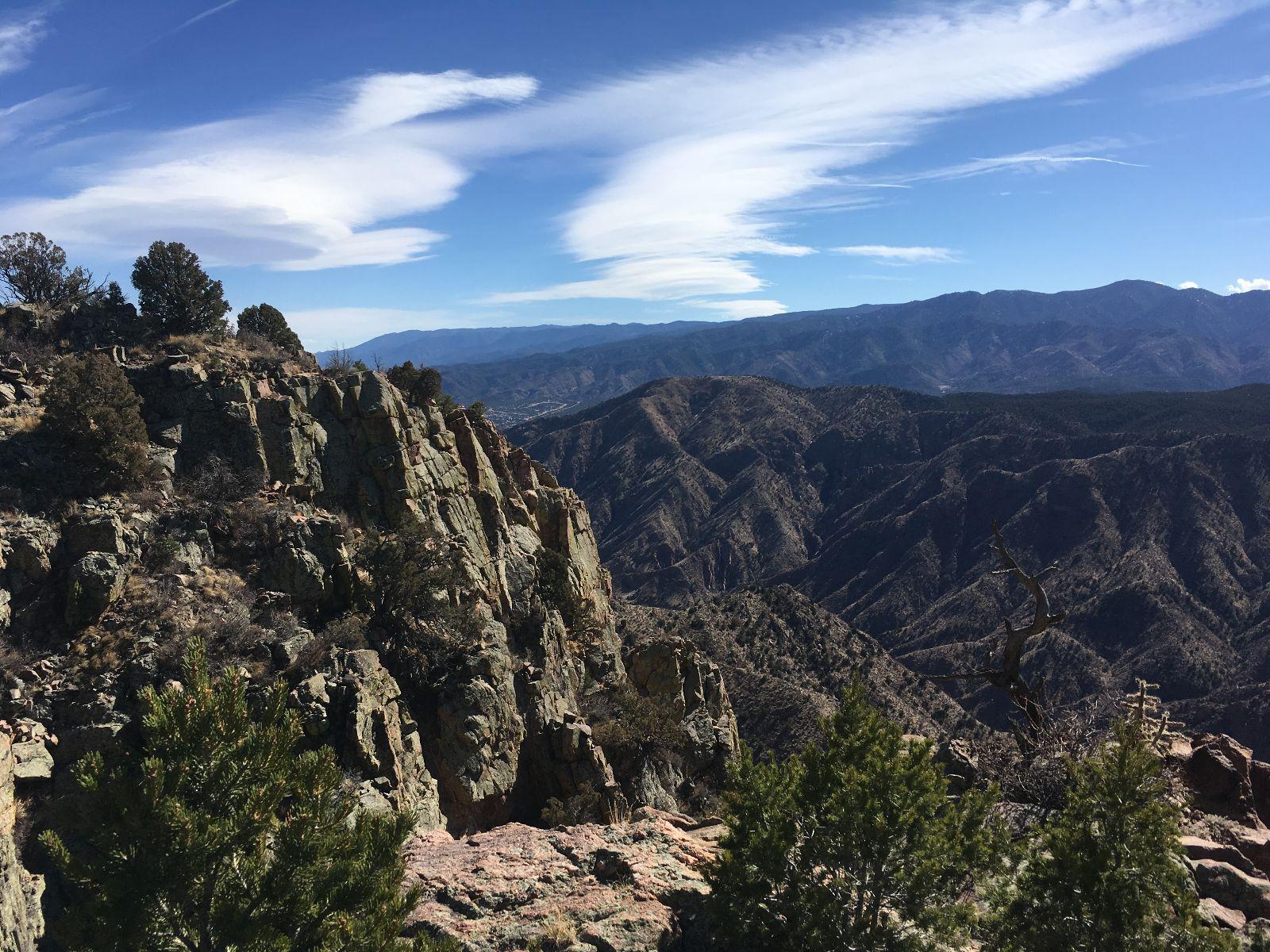 A stunning view of rugged mountains and valleys under a clear blue sky, featuring rocky cliffs in the foreground, sparse vegetation, and distant peaks. Royal Gorge Park Trail System mountain bike trail.