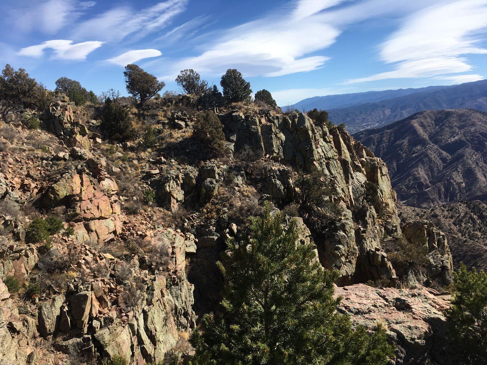 A rocky landscape with rugged terrain, featuring various shrubs and trees, under a clear blue sky with wispy clouds. The scene captures the undulating mountains in the background, creating a picturesque view of nature. Royal Gorge Park Trail System mountain bike trail.