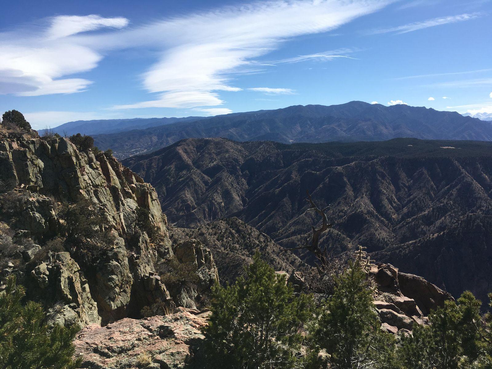 A scenic view of rugged mountain terrain under a clear blue sky, featuring rocky cliffs in the foreground and rolling hills in the background, with patches of trees dotting the landscape. Royal Gorge Park Trail System mountain bike trail.