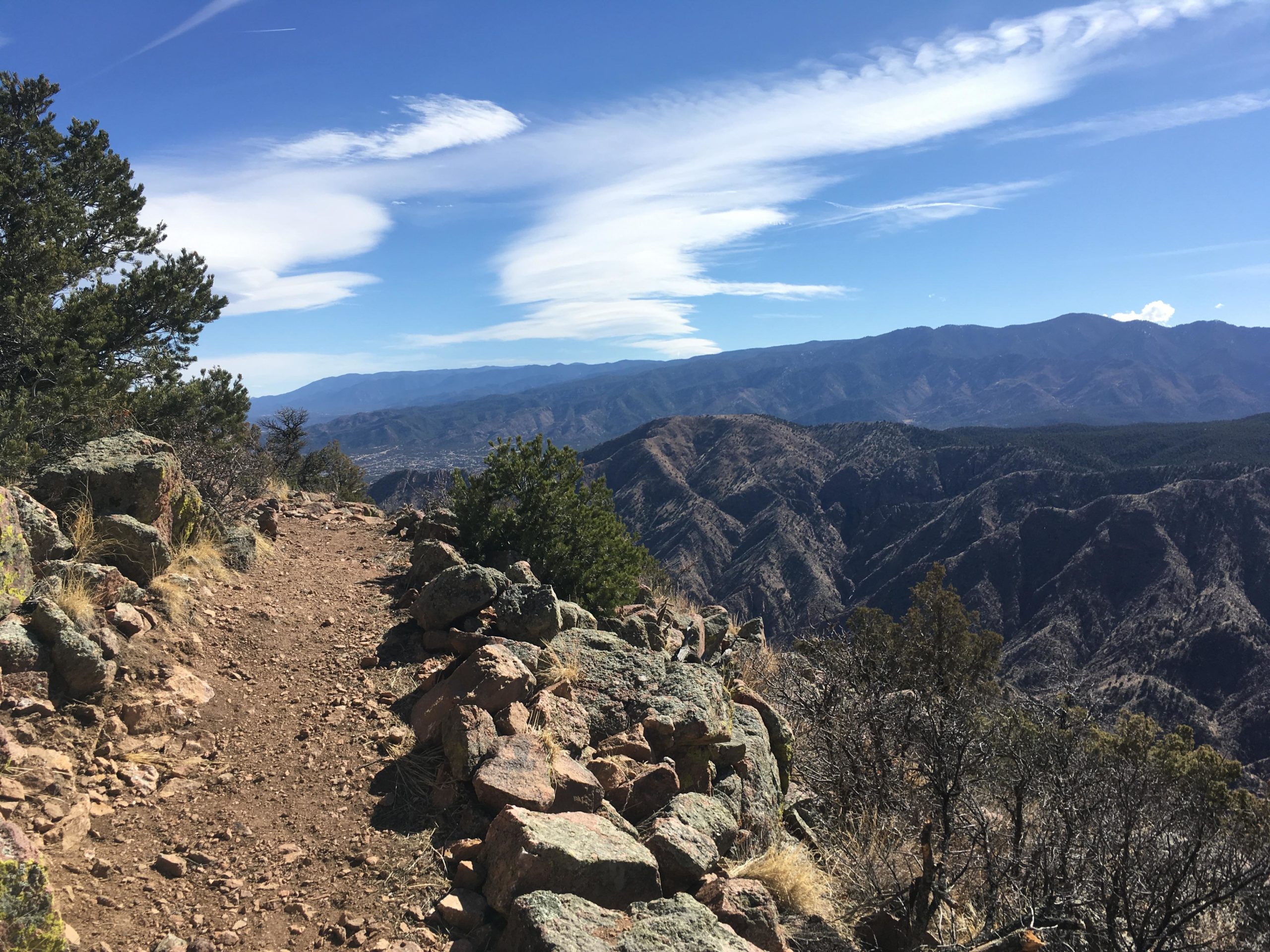 A rocky hiking trail along a mountain ridge, surrounded by trees and expansive mountain views under a blue sky with wispy clouds. Royal Gorge Park Trail System mountain bike trail.