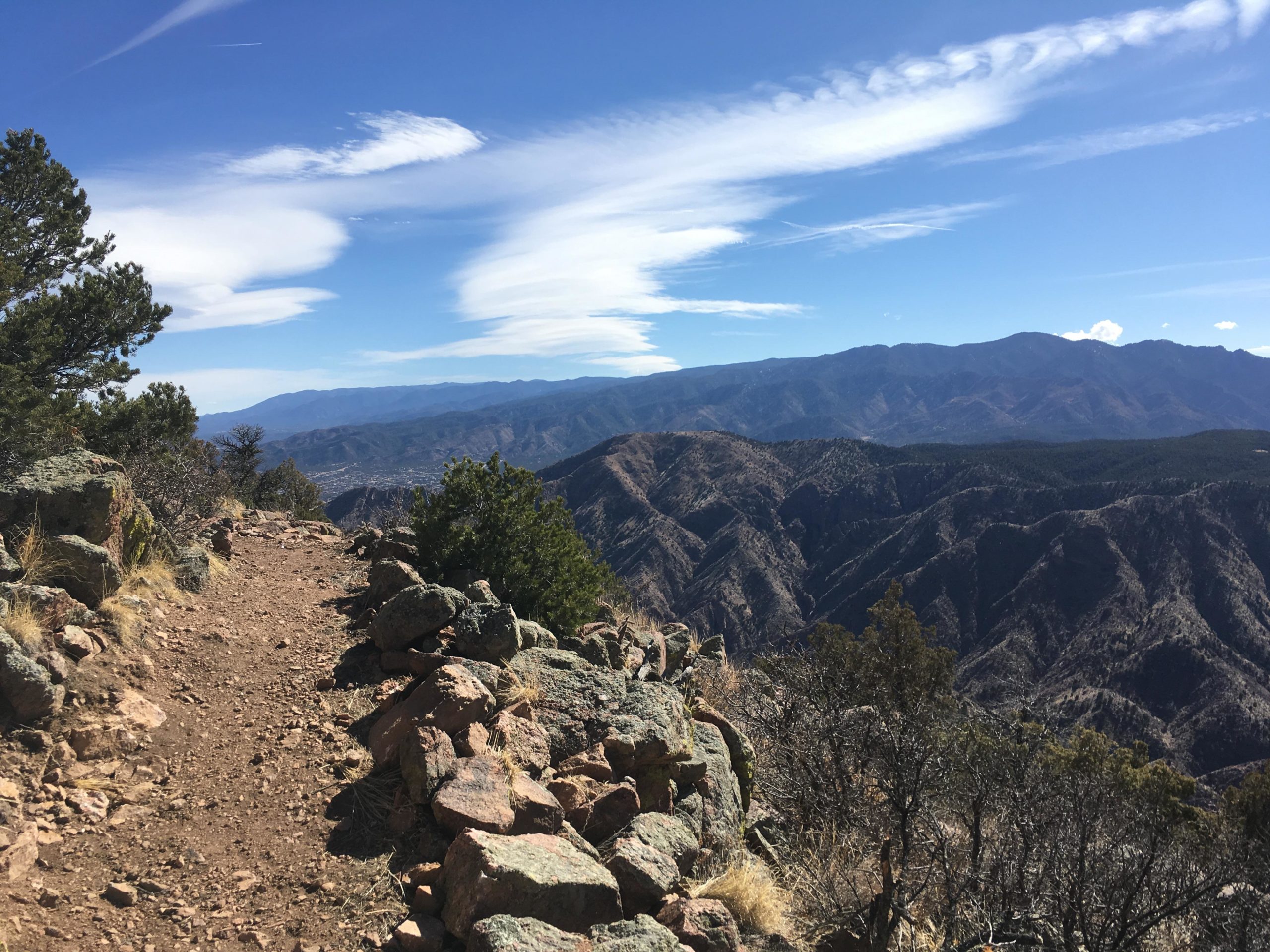 A scenic view of a mountain trail, bordered by rocky outcroppings and lush vegetation. The path winds along a ridgeline, offering expansive views of rolling mountains under a clear blue sky with wispy clouds. The landscape features a mix of rugged terrain and greenery, showcasing the beauty of nature. Royal Gorge Park Trail System mountain bike trail.