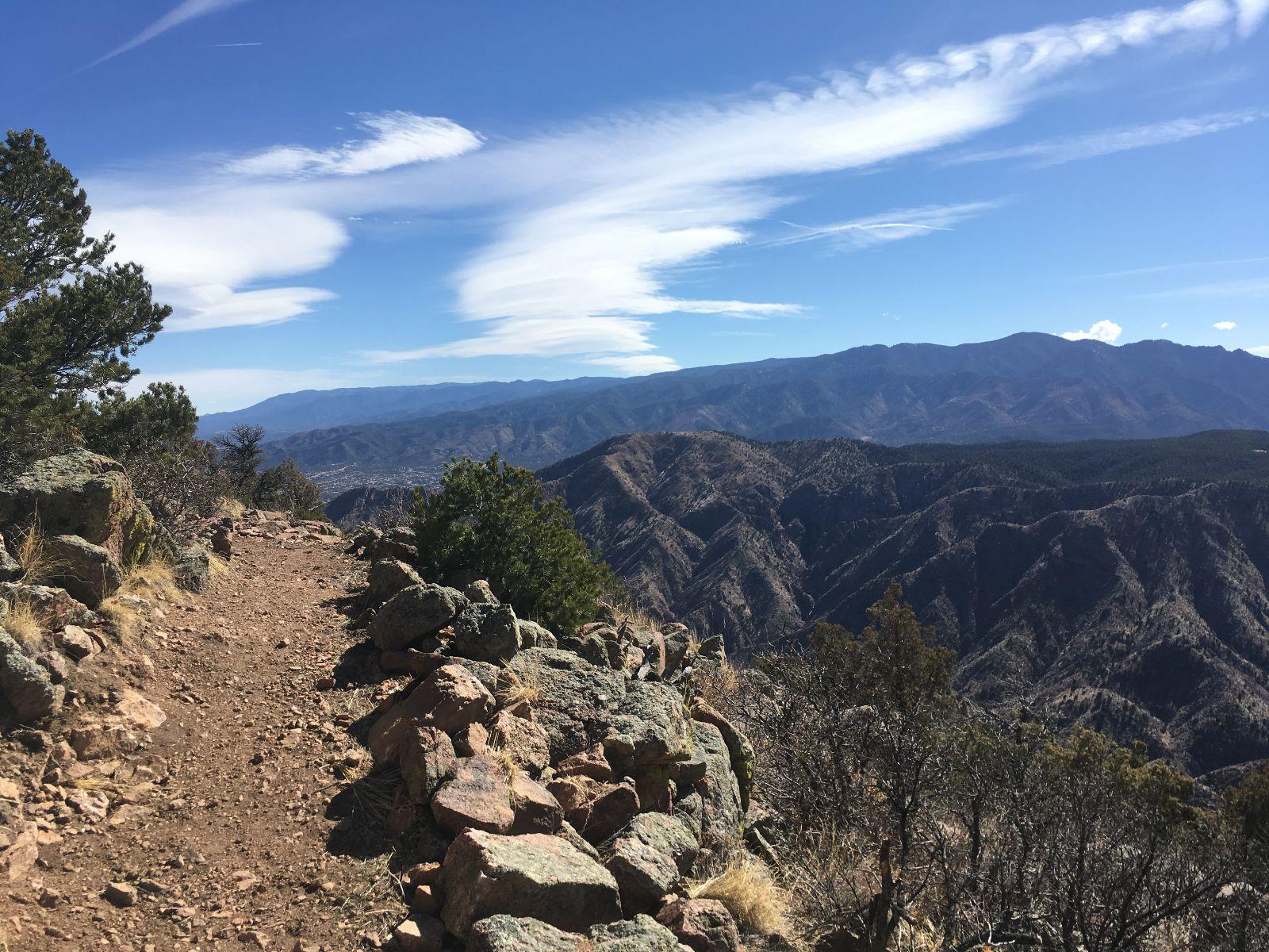 A scenic mountain trail winding along a rocky path, bordered by shrubs and trees. In the background, expansive mountain ranges are visible under a clear blue sky with wispy clouds. Royal Gorge Park Trail System mountain bike trail.