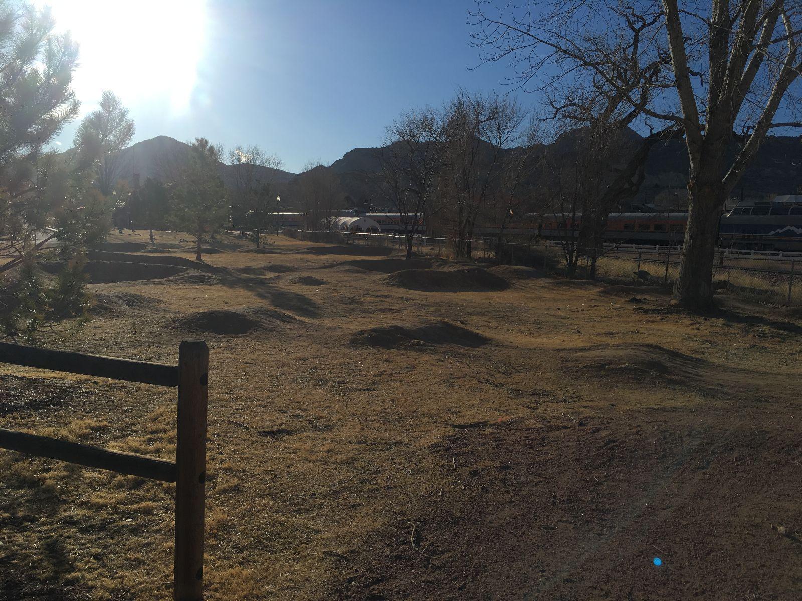 A sunlit outdoor scene featuring a grassy area with several small mounds, surrounded by sparse trees and mountains in the background. A wooden fence is visible in the foreground, and a train can be seen in the distance behind a fence. The atmosphere is clear and bright, suggesting a pleasant daytime setting. Royal Gorge Park Trail System mountain bike trail.