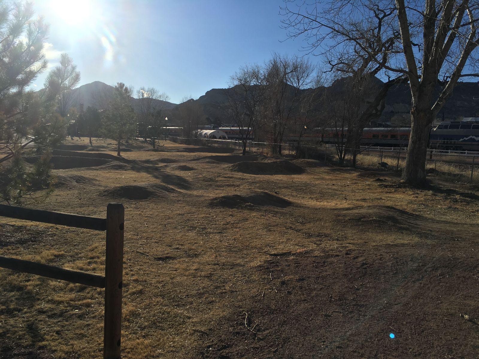 A sunlit landscape featuring rolling hills in a grassy area, with sparse trees and mountains in the background. A wooden fence is in the foreground, and a train can be seen in the distance along a track. The scene conveys a serene outdoor environment. Royal Gorge Park Trail System mountain bike trail.