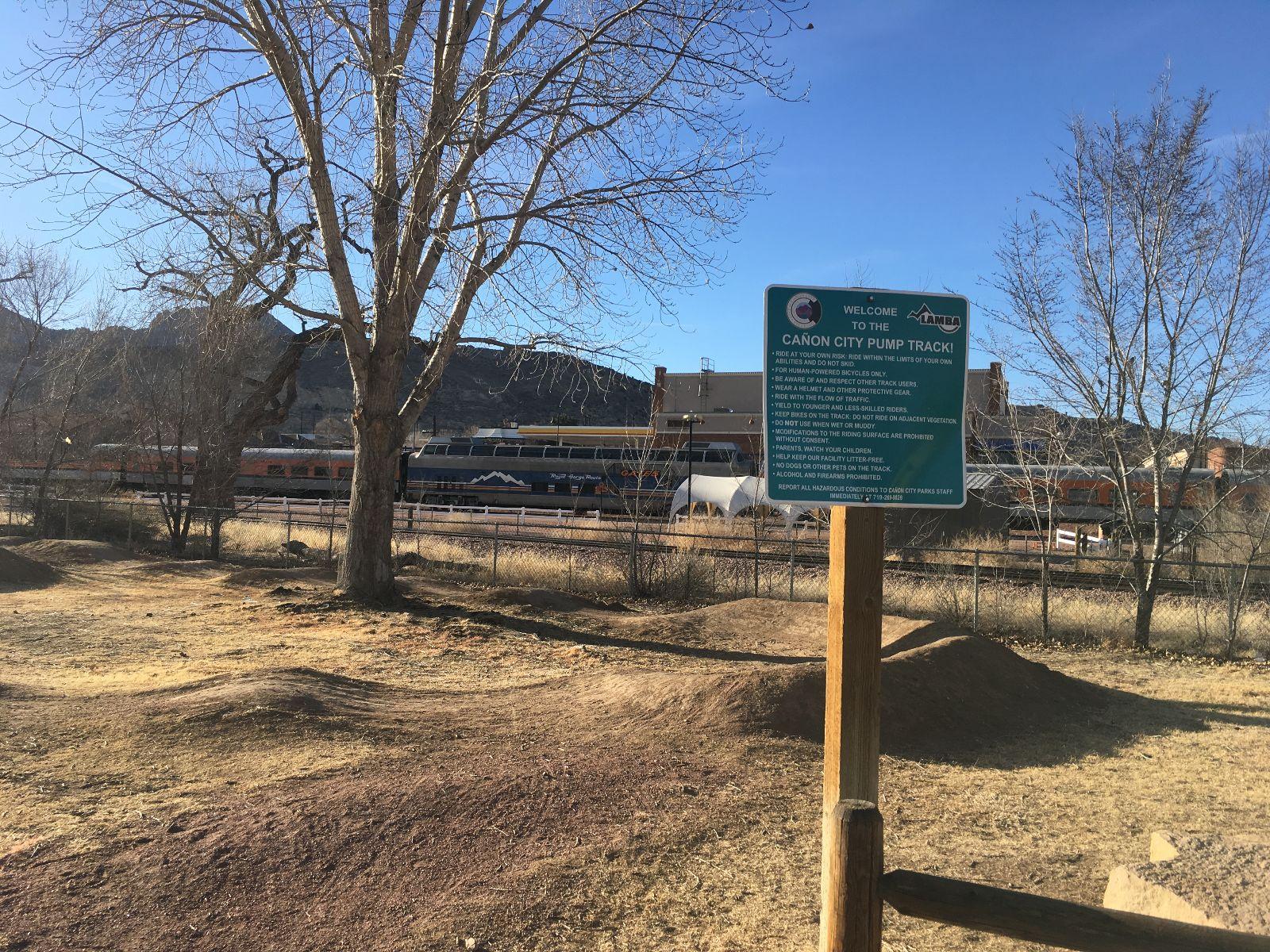 A view of a tree and grassy area in front of a sign that reads "Welcome to the Canon City Pump Track!" The background features a train passing by in front of a mountain landscape under a clear blue sky. The sign provides rules and guidelines for using the pump track. Royal Gorge Park Trail System mountain bike trail.