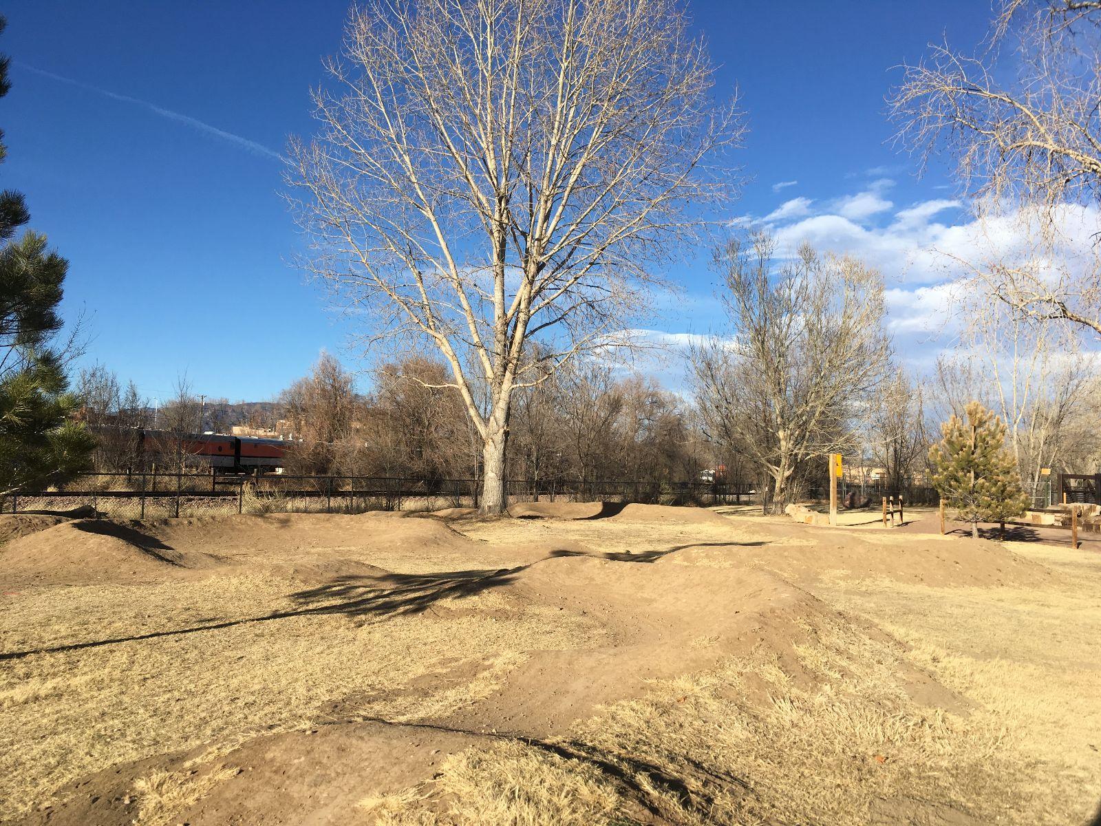 A landscape view of a park featuring brown dirt mounds and patches of dry grass under a clear blue sky. Leafless trees are scattered throughout the area, with a glimpse of a fenced playground in the background. The scene suggests a peaceful outdoor space. Royal Gorge Park Trail System mountain bike trail.