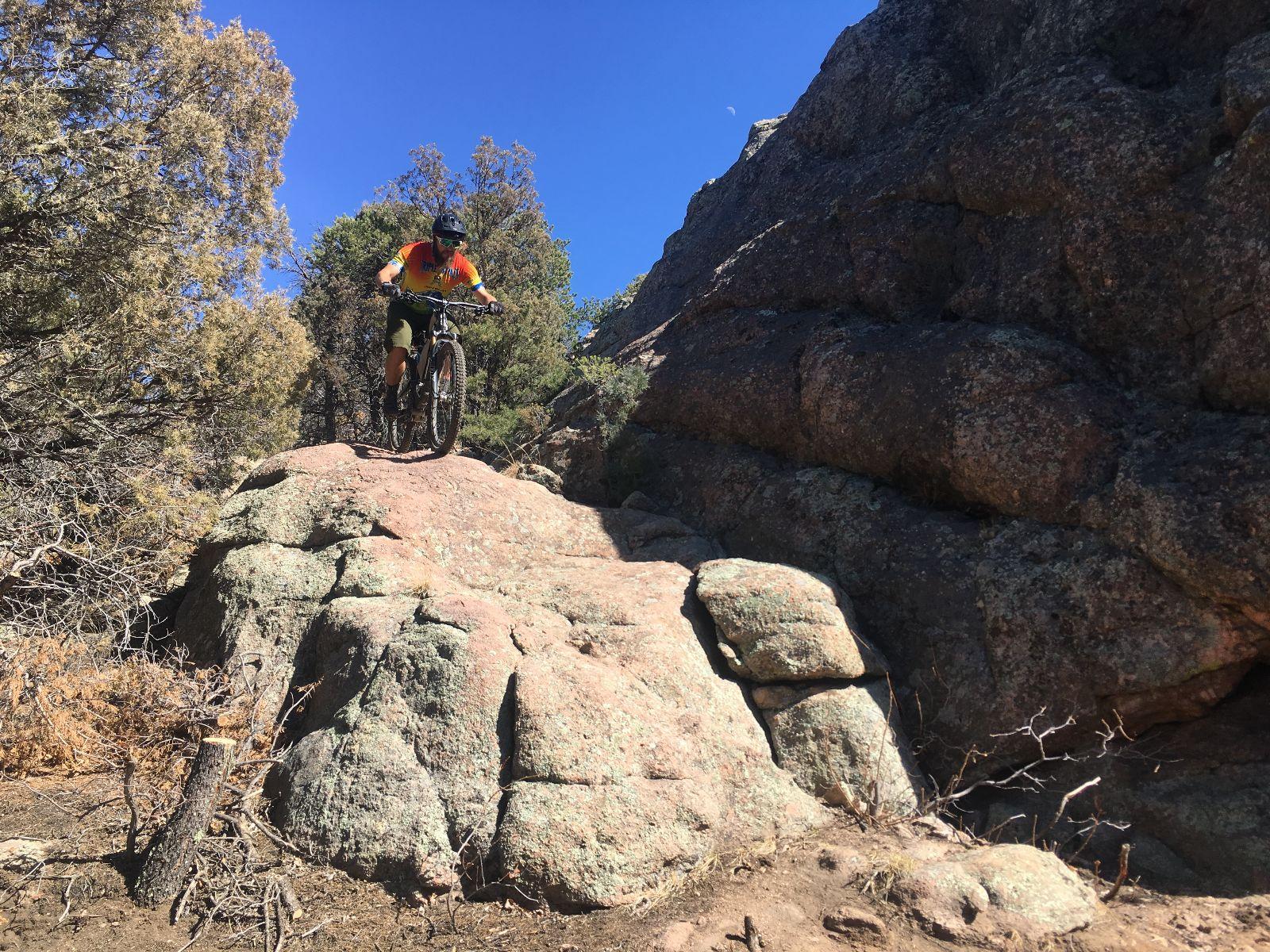 A mountain biker navigating a rocky trail, balancing on large boulders surrounded by shrubs and trees under a clear blue sky. Royal Gorge Park Trail System mountain bike trail.