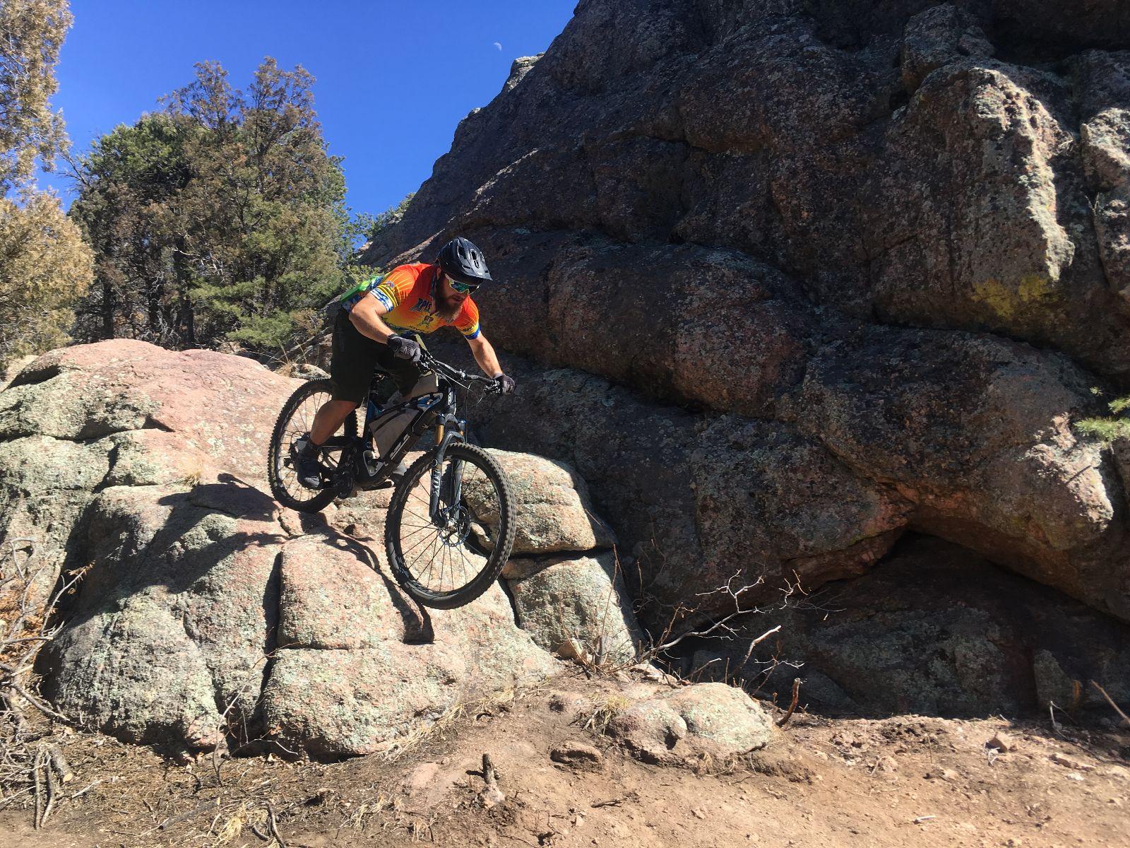 A mountain biker in a colorful jersey descends from a rocky outcrop, navigating the uneven terrain amidst trees and a clear blue sky. Royal Gorge Park Trail System mountain bike trail.