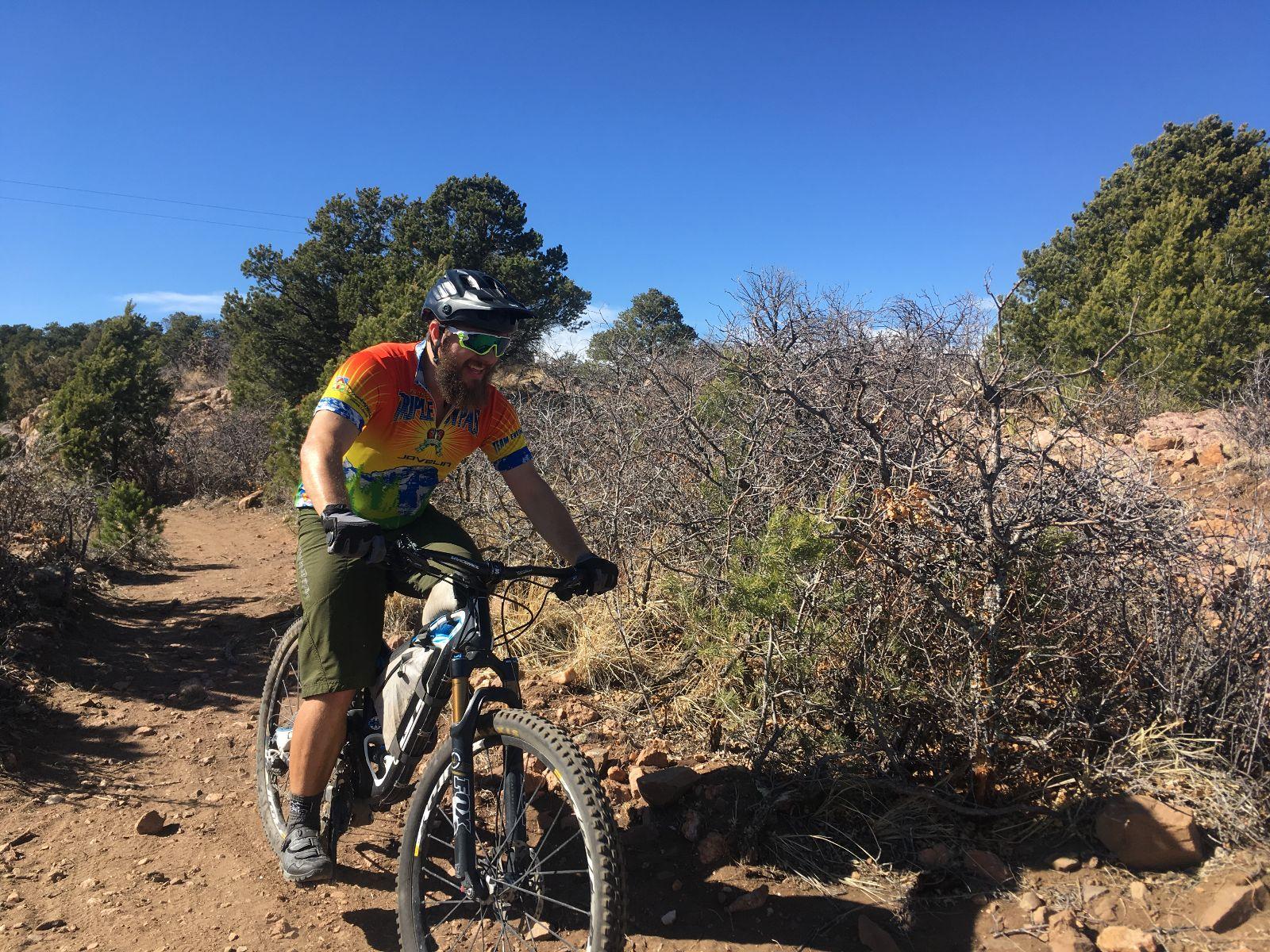 A mountain biker riding on a dirt trail surrounded by sparse vegetation under a clear blue sky. The rider wears a colorful cycling jersey and helmet, smiling as he navigates the path. Royal Gorge Park Trail System mountain bike trail.