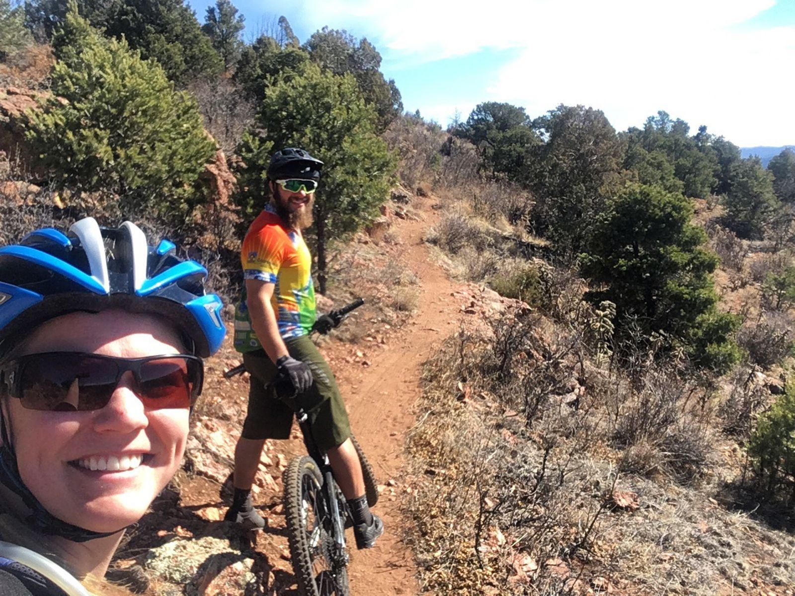 Two mountain bikers smiling for a selfie on a trail surrounded by trees and rocky terrain. One rider is in the foreground wearing a blue helmet and sunglasses, while the other is in the background wearing a colorful jersey and standing next to their bike. A clear sky can be seen above, indicating a sunny day. Royal Gorge Park Trail System mountain bike trail.