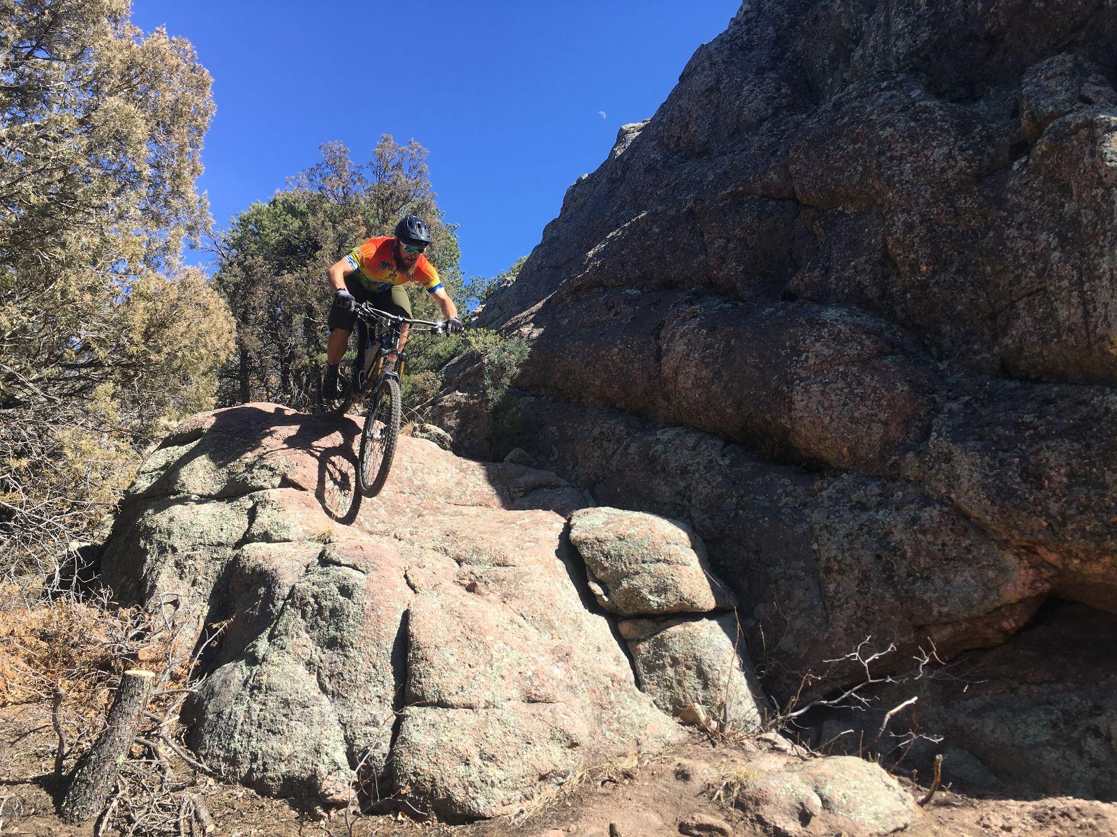 A mountain biker in a brightly colored jersey rides down a rocky terrain, navigating over large boulders against a clear blue sky. Surrounding vegetation includes sparse trees, adding to the rugged outdoor landscape. Royal Gorge Park Trail System mountain bike trail.