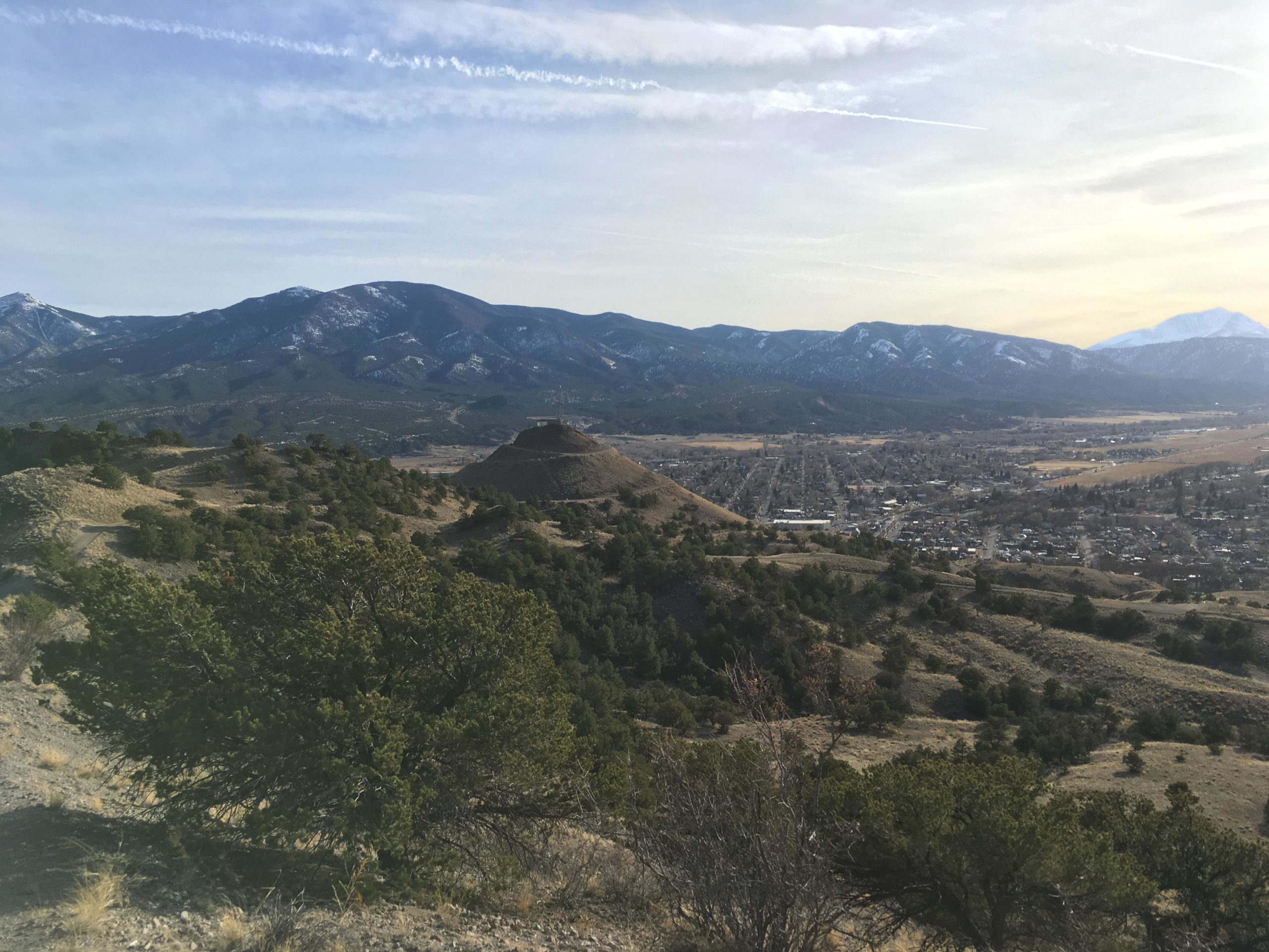 A scenic view of a mountainous landscape featuring rolling hills, a valley with a small town, and snow-capped peaks in the background. The foreground includes greenery and trees, while the sky is partly cloudy, creating a tranquil atmosphere. Arkansas Hills mountain bike trail.