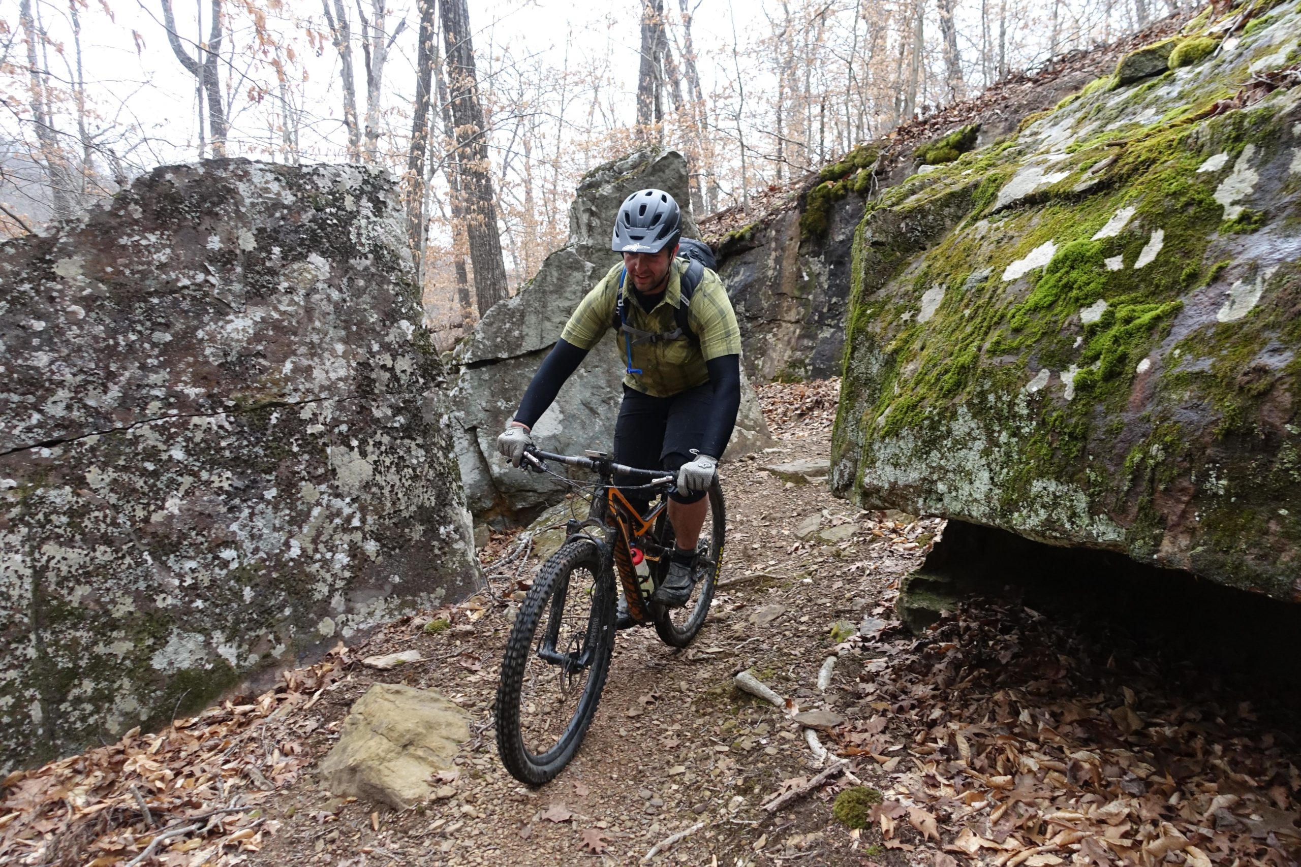 A mountain biker navigating a narrow trail surrounded by large rocks and trees in a forested area. The cyclist is wearing a helmet and biking gear, focused on maneuvering the terrain. Fallen leaves and moss are visible on the ground. Upper Buffalo Headwaters Trail System mountain bike trail.