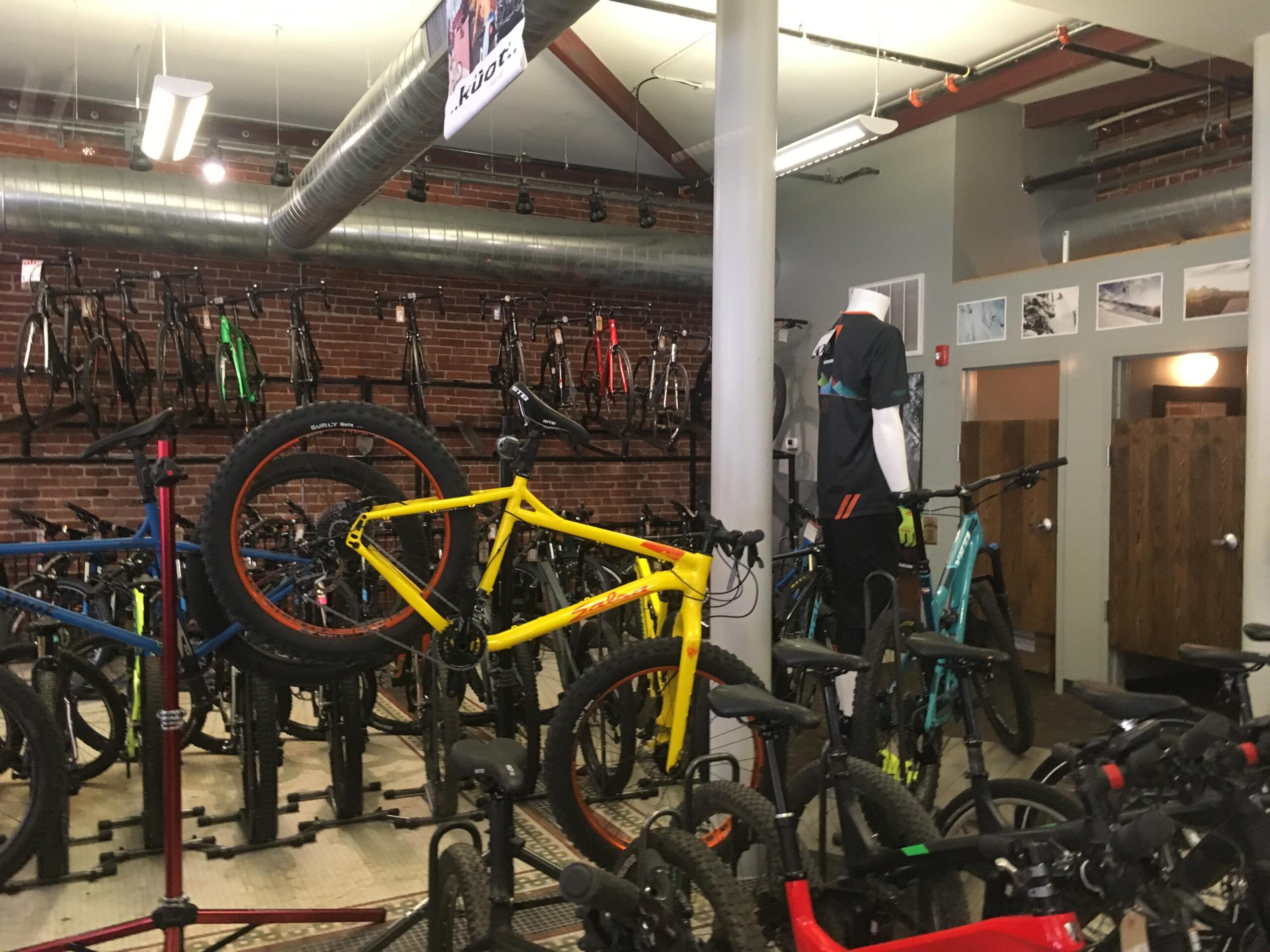 A vibrant bicycle shop interior featuring a variety of bikes on display. Brightly colored bikes, including a yellow one, are prominently positioned amidst a backdrop of brick walls. The shop includes a mannequin dressed in cycling attire and additional bikes hanging on racks in the background. The setting is lively and inviting for cycling enthusiasts.