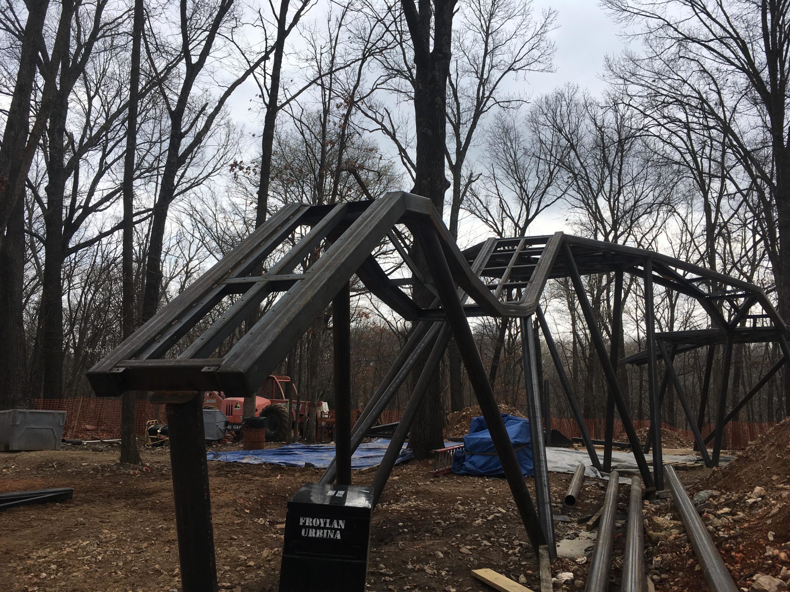 An unfinished metal structure resembling a roller coaster, surrounded by bare trees and construction equipment. The ground is covered with dirt and debris, and there are blue tarps and a black box with the name "Froylan Urbina" visible in the foreground. The sky is overcast, indicating a cloudy day. Coler Mountain Bike Preserve mountain bike trail.