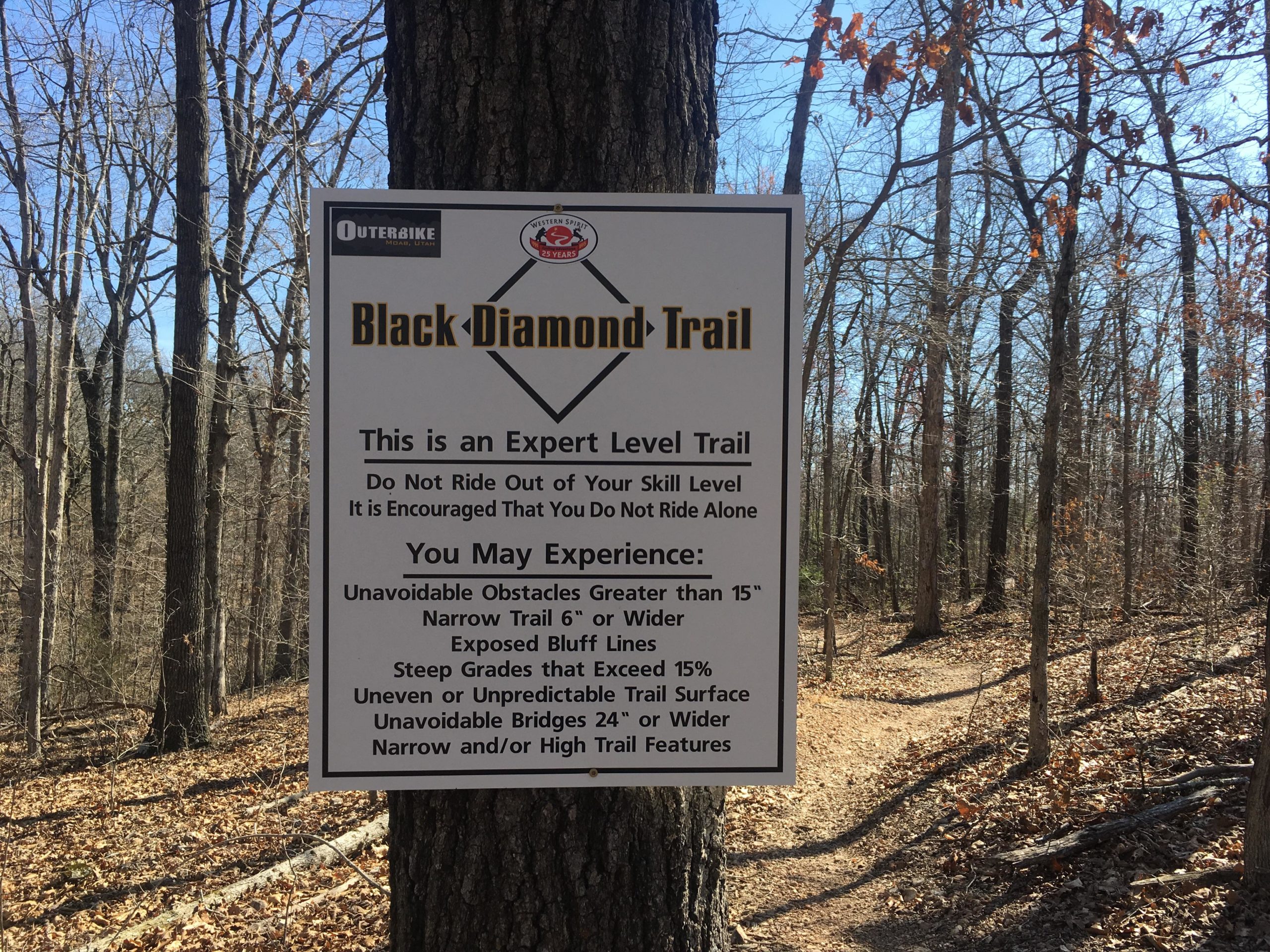 Sign marking the Black Diamond Trail, indicating it is an expert-level trail. The sign lists safety advisories, encouraging riders not to exceed their skill level and to ride with others. It highlights possible experiences such as unavoidable obstacles, narrow trails, steep grades, and other challenging features. Surrounding landscape includes trees and a clear blue sky. Back 40 mountain bike trail.