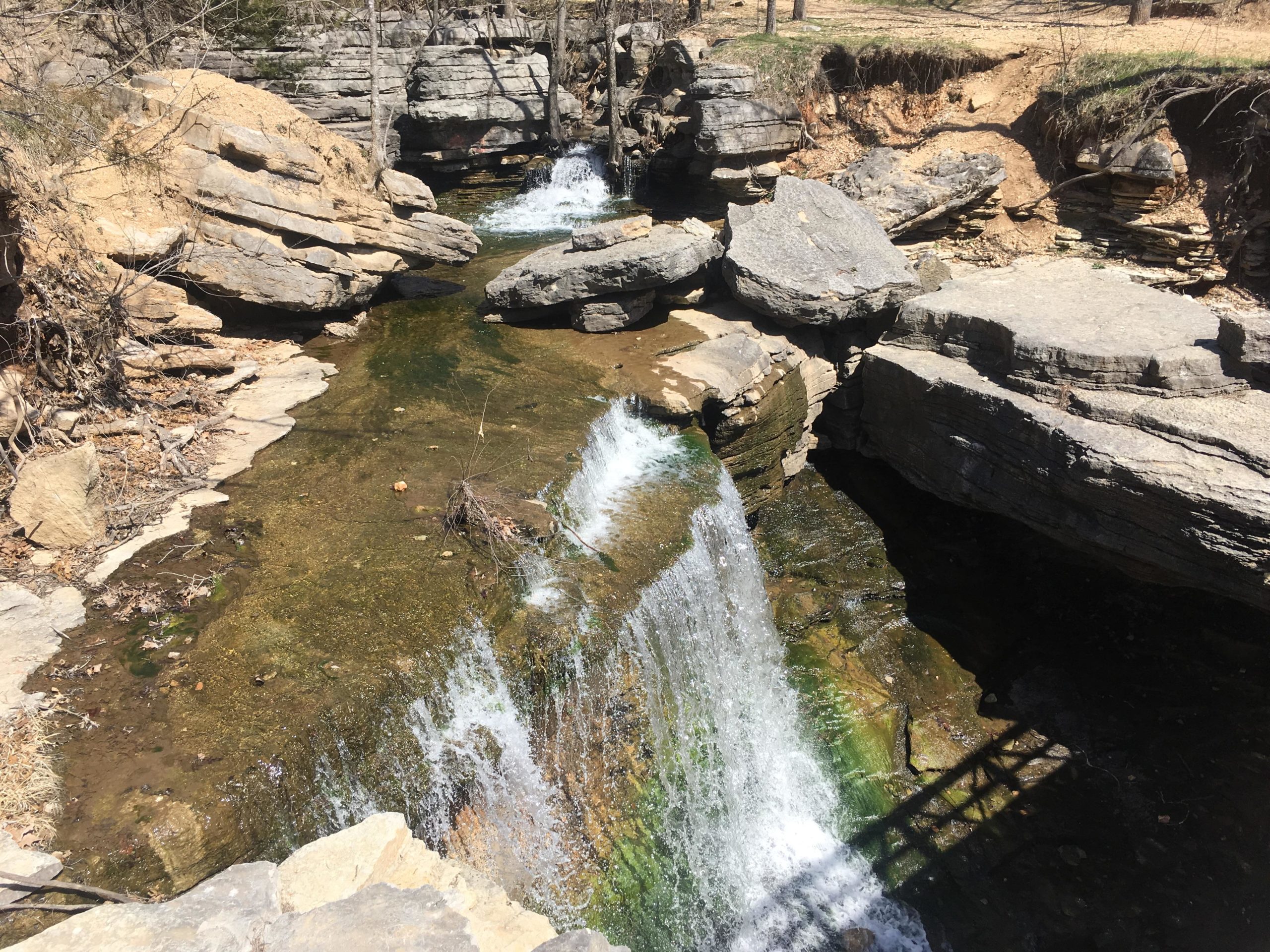A natural stream flowing over rocky terrain, with small waterfalls cascading into clear water. Surrounding the stream are large boulders and patches of dry grass, under a bright, sunny sky. Back 40 mountain bike trail.