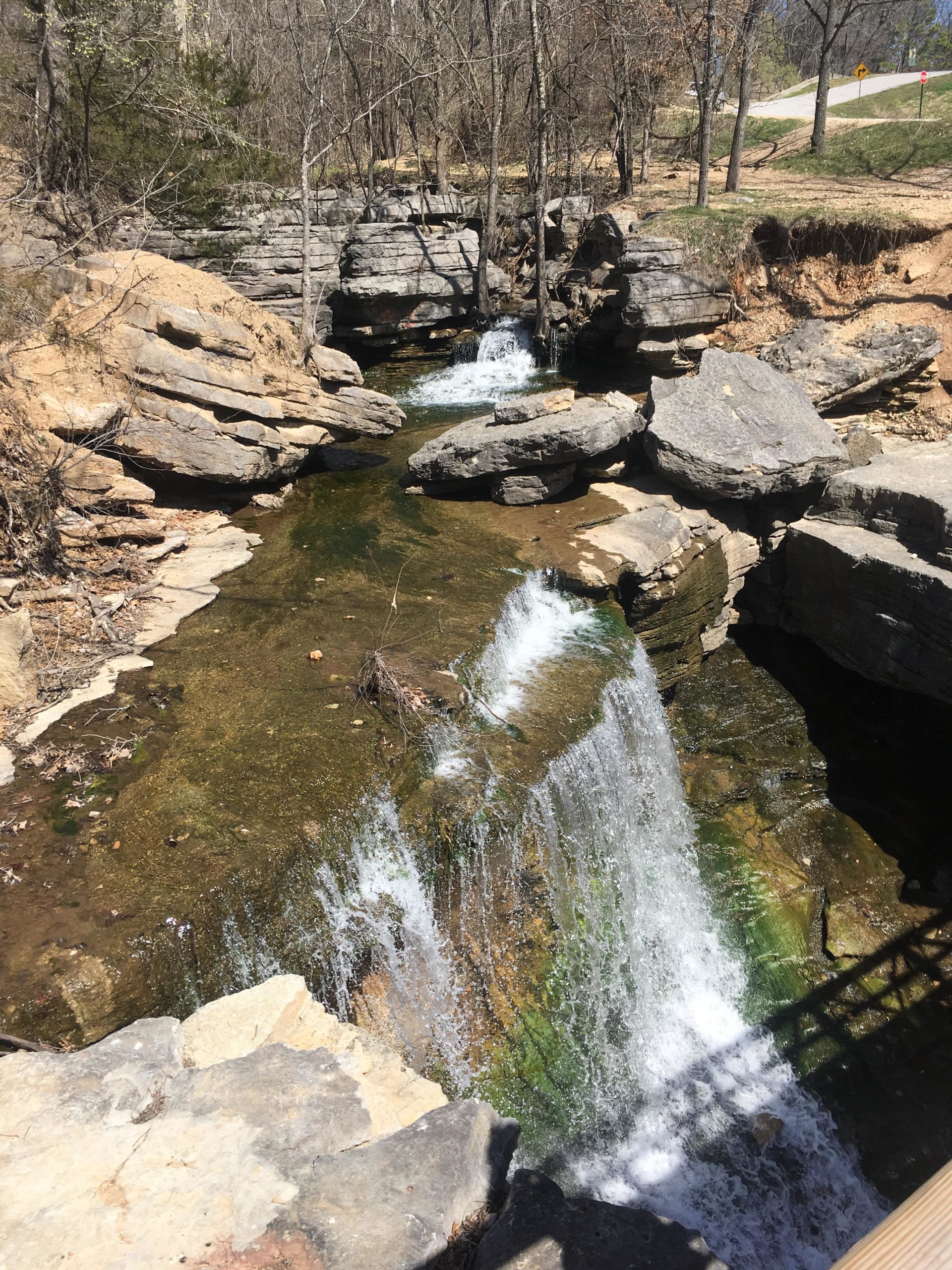 A serene natural scene featuring a small waterfall cascading over rocky ledges into a clear stream below. Surrounding the water are scattered rocks and sparse trees, suggesting a tranquil outdoor environment. The sunlight illuminates the water and highlights the texture of the stones, creating a peaceful atmosphere. Back 40 mountain bike trail.