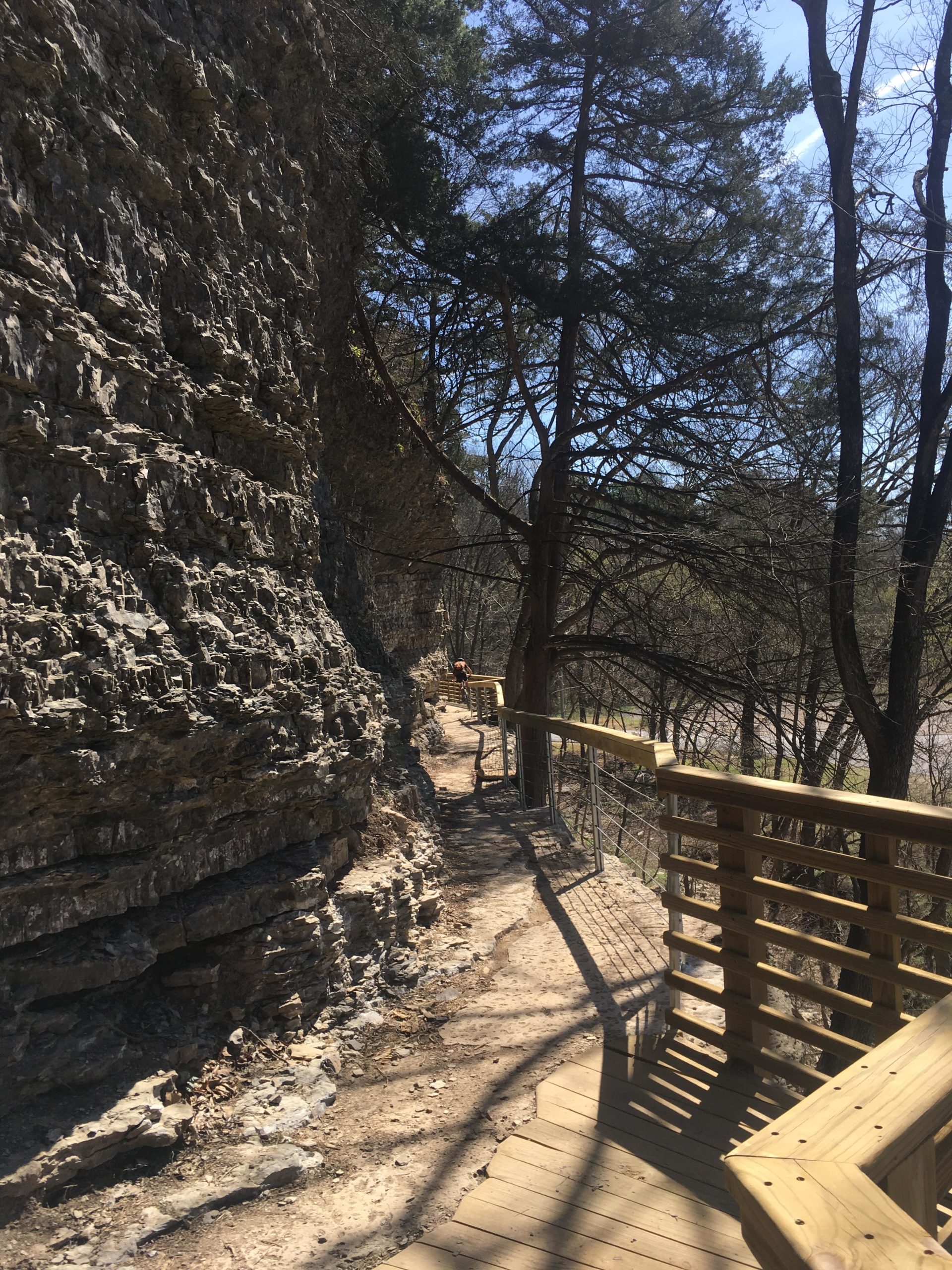 A wooden boardwalk meanders alongside a rocky cliff, surrounded by trees under a clear blue sky. The path, bordered by a railing, offers a peaceful outdoor setting perfect for hiking. Back 40 mountain bike trail.
